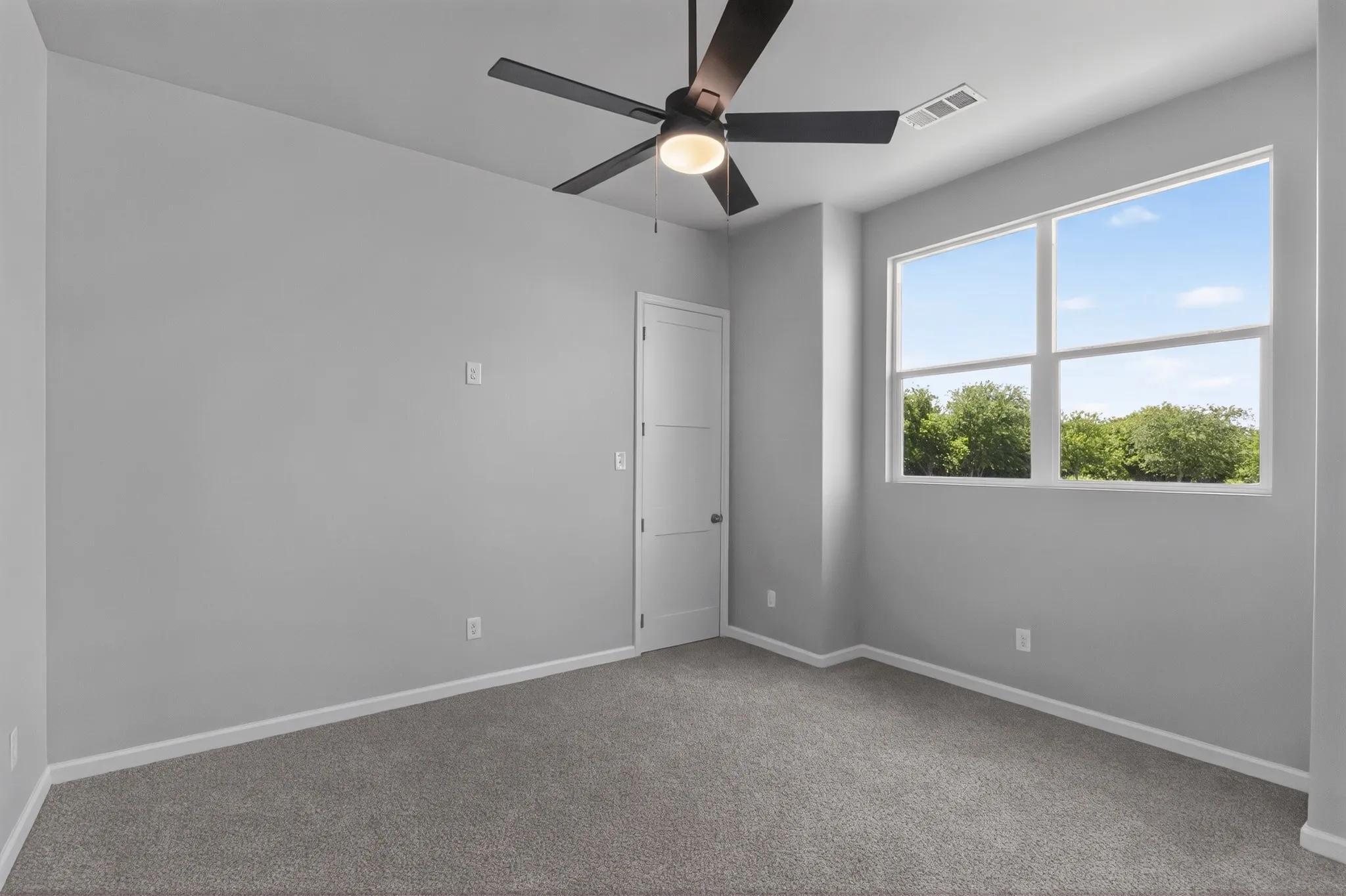 Spacious room featuring plush gray carpet, a modern ceiling fan with integrated lighting, and a large multi-pane window showcasing green tree foliage against a blue sky