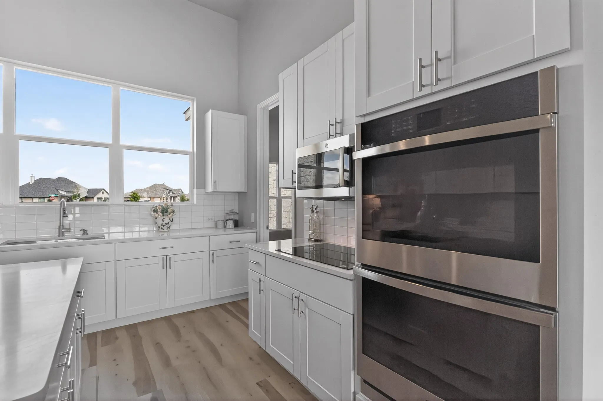 Kitchen featuring white shaker cabinetry, stainless steel double ovens, wood-finish flooring, a large window, and subway tile backsplash