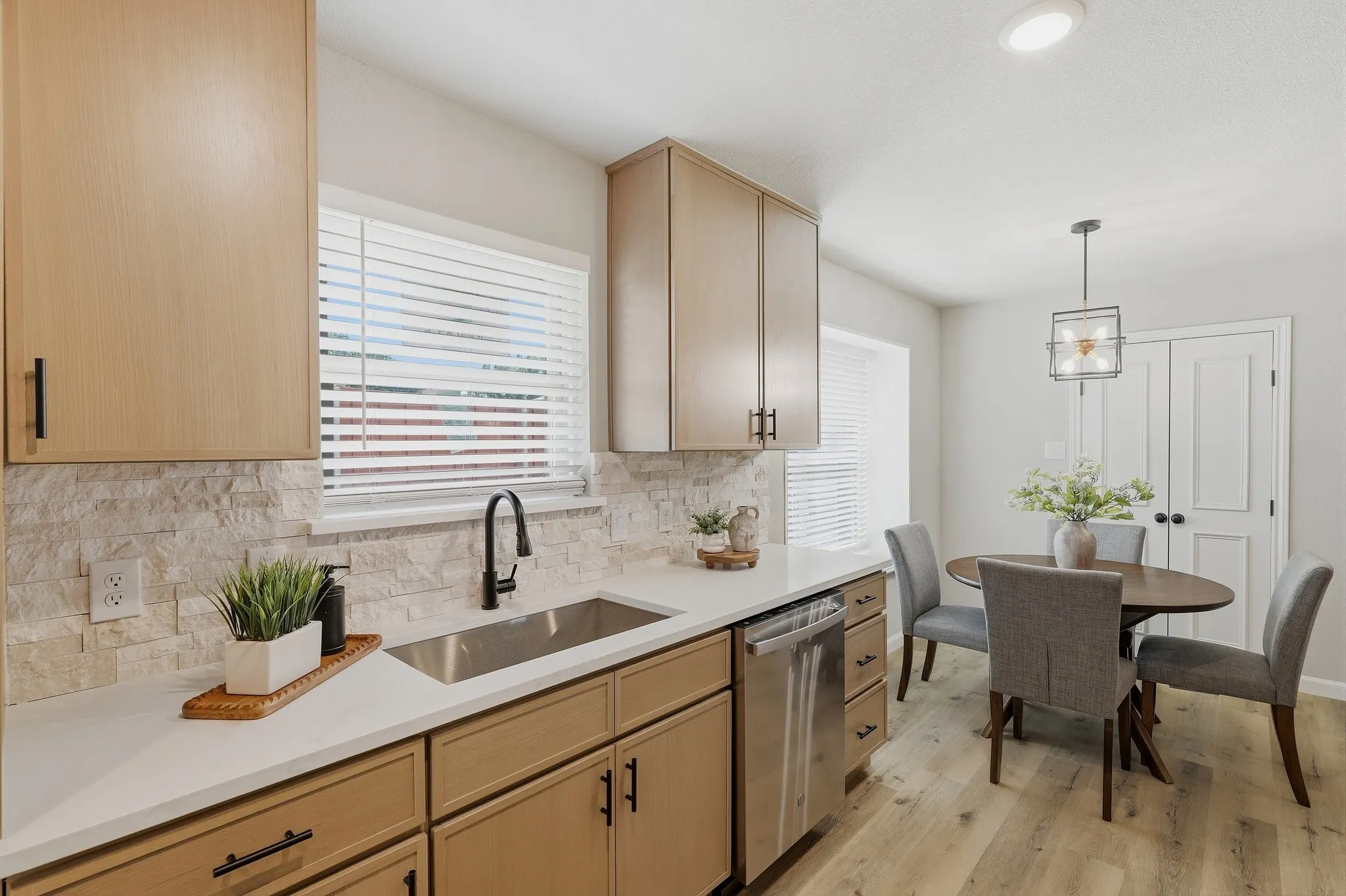 Light wood-finish flooring, light wood cabinetry with dark hardware, stainless steel sink with matte black faucet, light stone tile backsplash, and recessed lighting