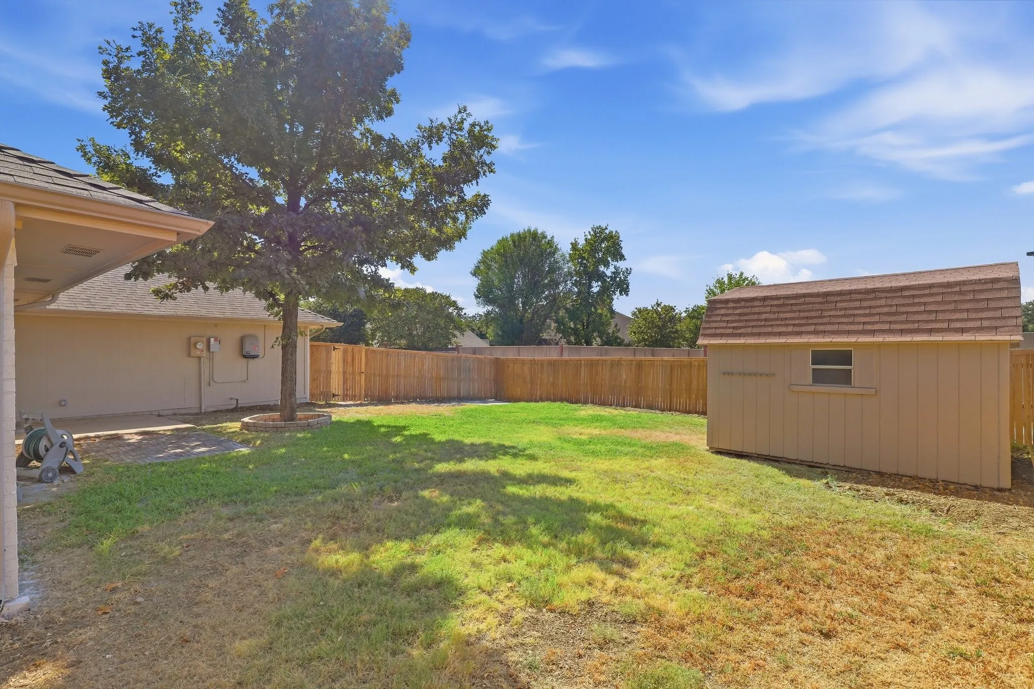 Fenced backyard featuring a storage shed with shingled roof, mature tree, and concrete patio