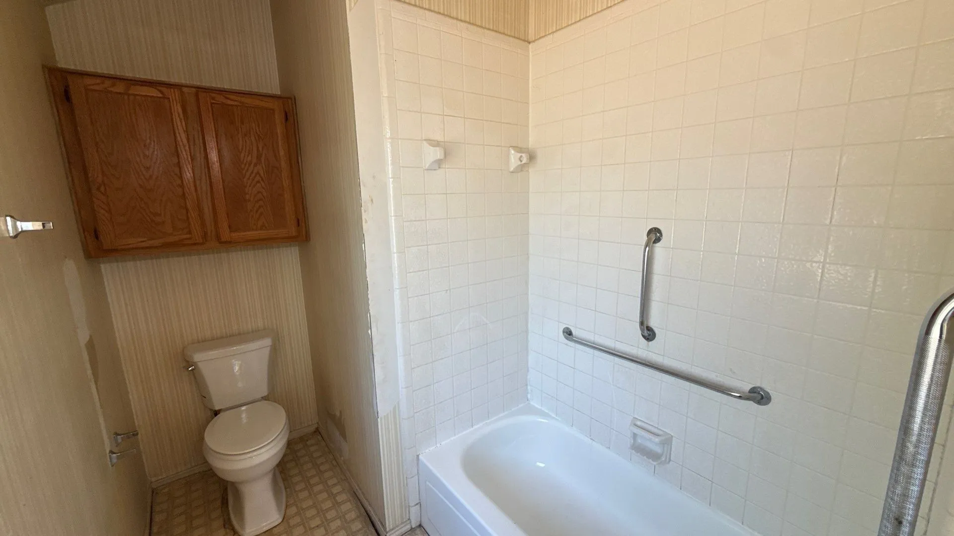White ceramic tile shower surround with integrated grab bars, a white bathtub, and wood-finish wall cabinetry