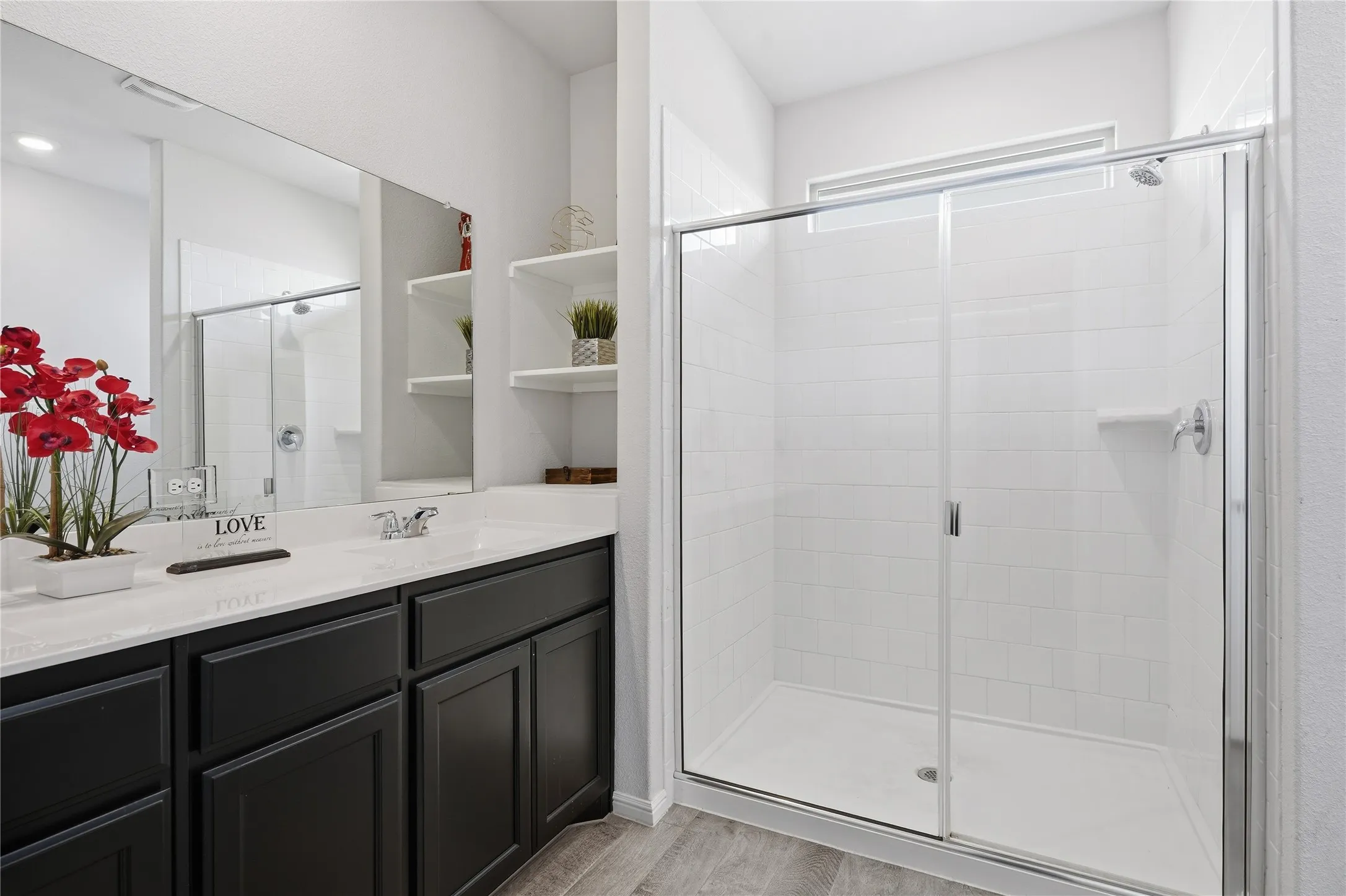 Bathroom featuring a dark-finish vanity with a light-toned countertop, large wall-mounted mirror, walk-in shower with white tile surround and sliding glass doors, and built-in shelving