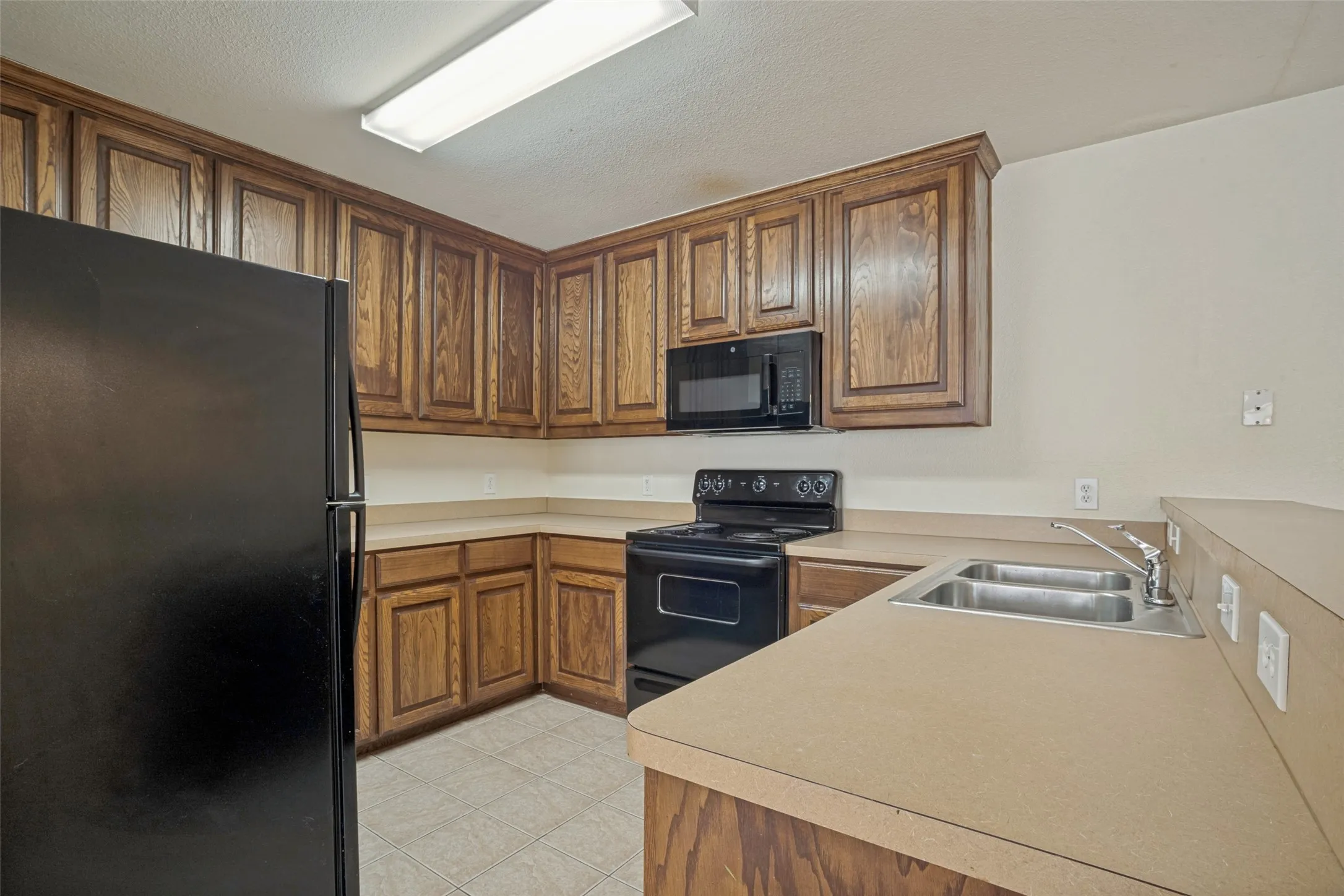 Kitchen featuring wood cabinetry, black appliances, a double basin stainless steel sink, and light-toned countertops