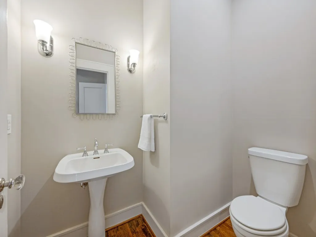 Pedestal sink with chrome fixtures, mirror with decorative frame, wall-mounted sconces, and wood-finish flooring