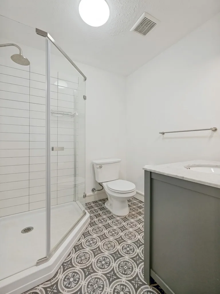 Walk-in shower with clear glass enclosure and subway tile surround, patterned floor tiling, single vanity with light-toned countertop, and modern lighting