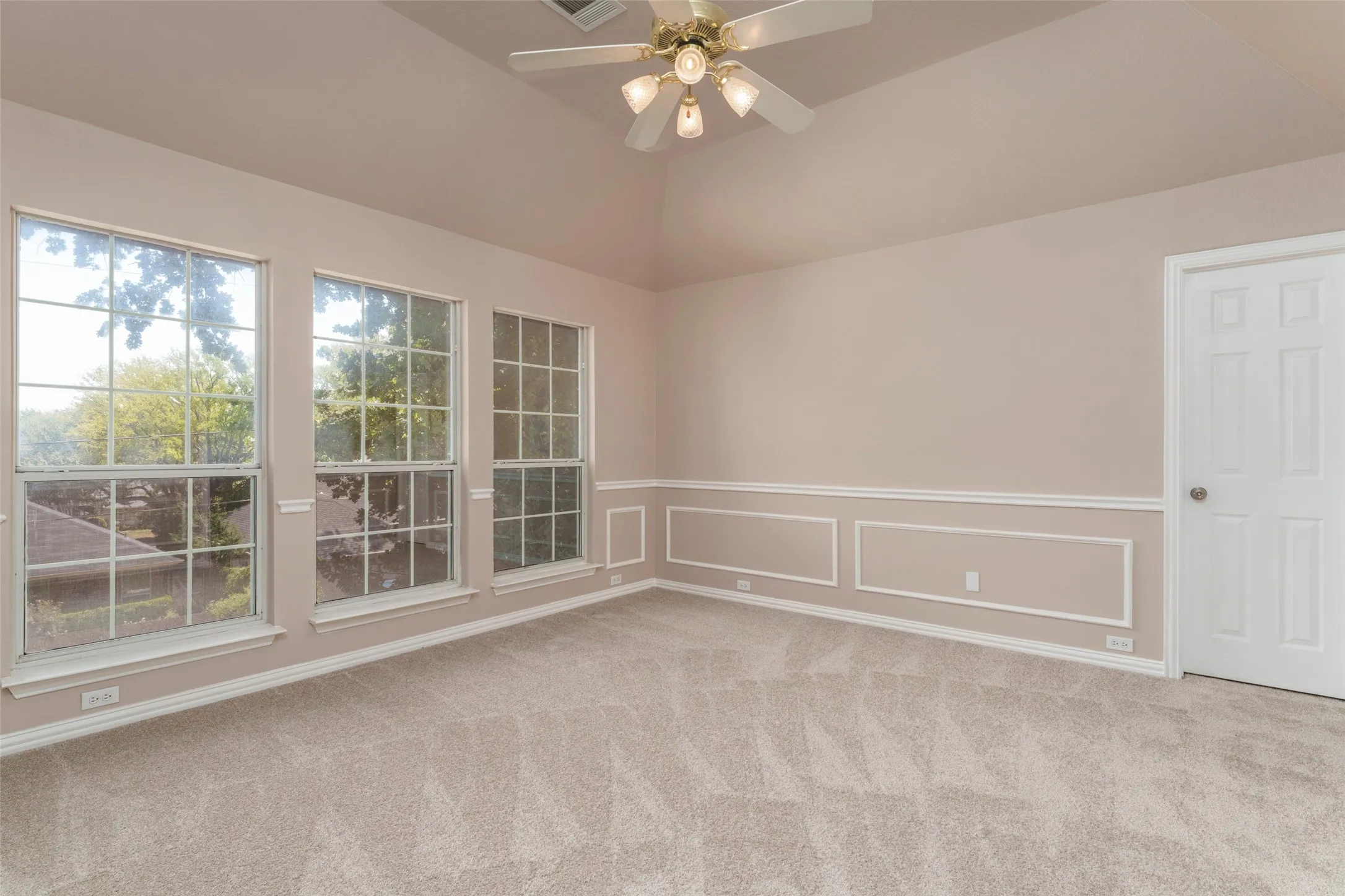 Vaulted ceiling with an attached ceiling fan and light fixture, three large windows, wainscoting with decorative trim, and neutral-toned carpeting