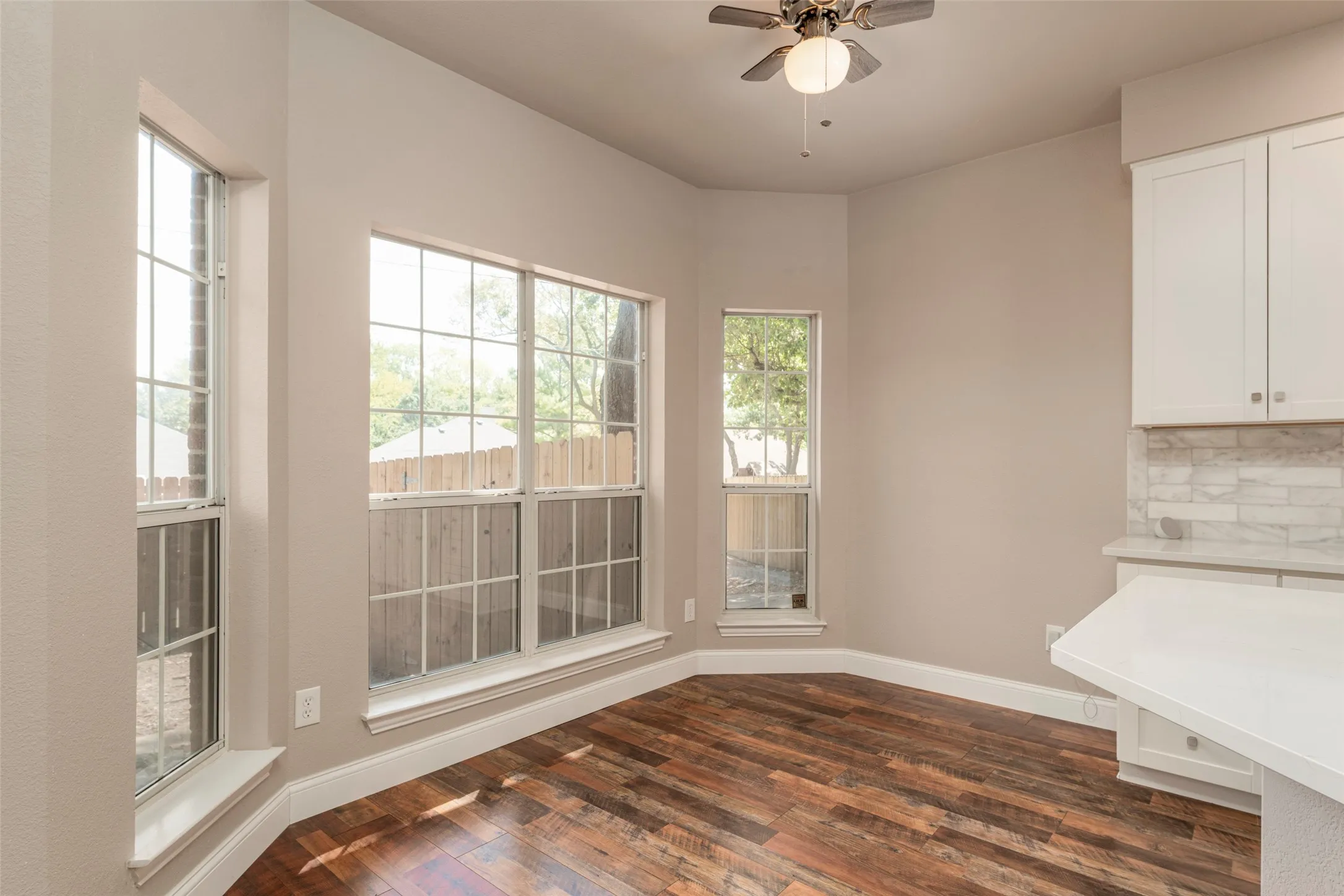 Wood-finish flooring, multiple windows with grid patterns, ceiling fan with integrated lighting, white upper cabinetry, and a light-toned backsplash