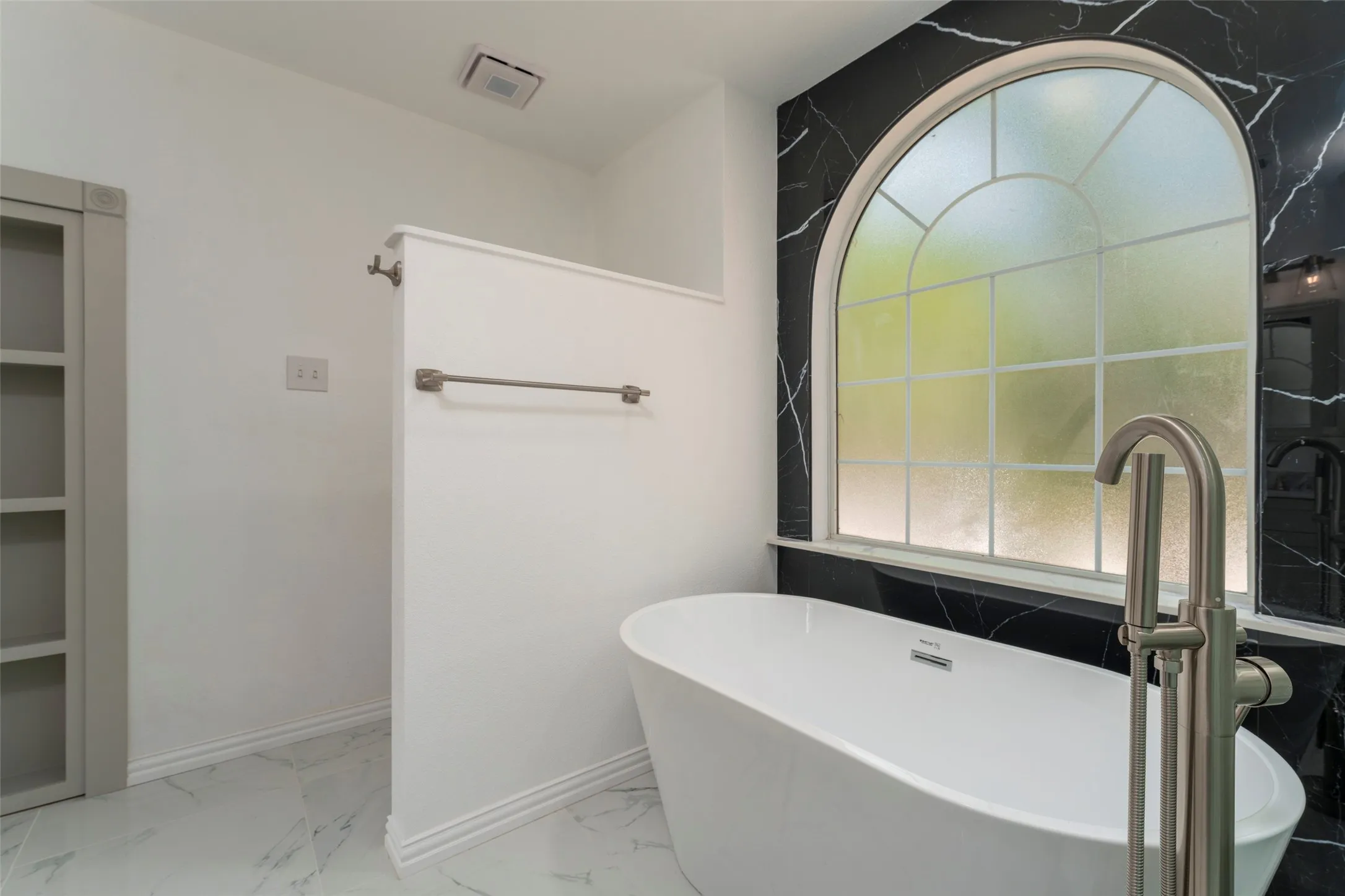 Freestanding soaking tub with floor-mounted brushed nickel faucet, large arched window, black and white veined wall paneling, built-in shelving, and white marble-look flooring