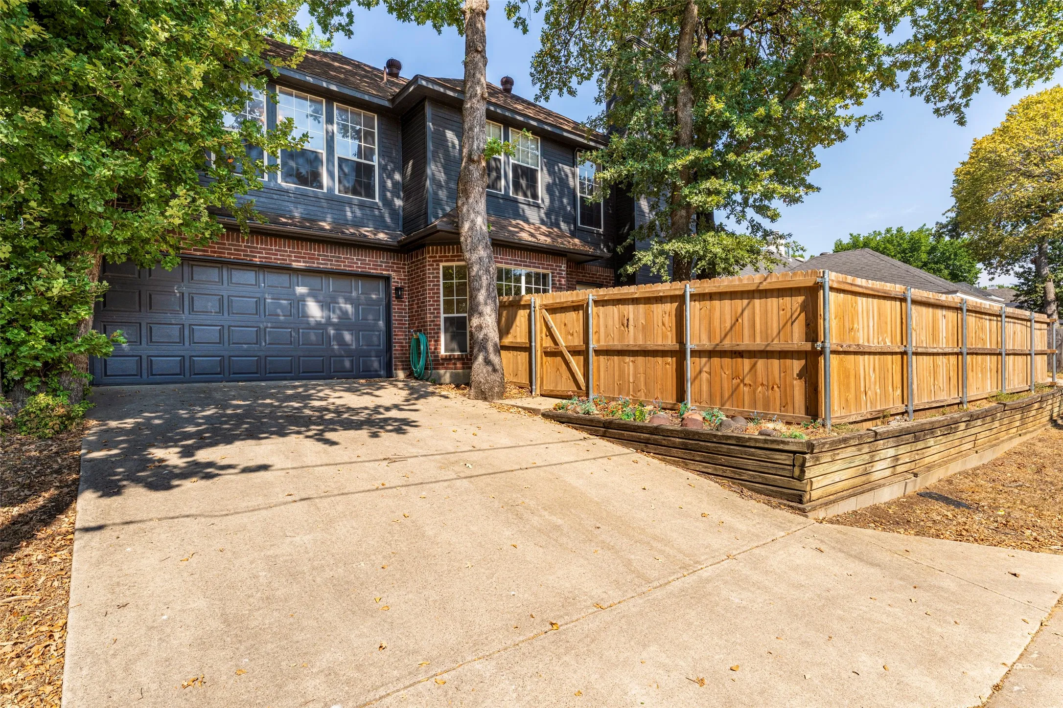 Two-story exterior with brick and siding facade, attached two-car garage, concrete driveway, wood privacy fence, and raised planter bed