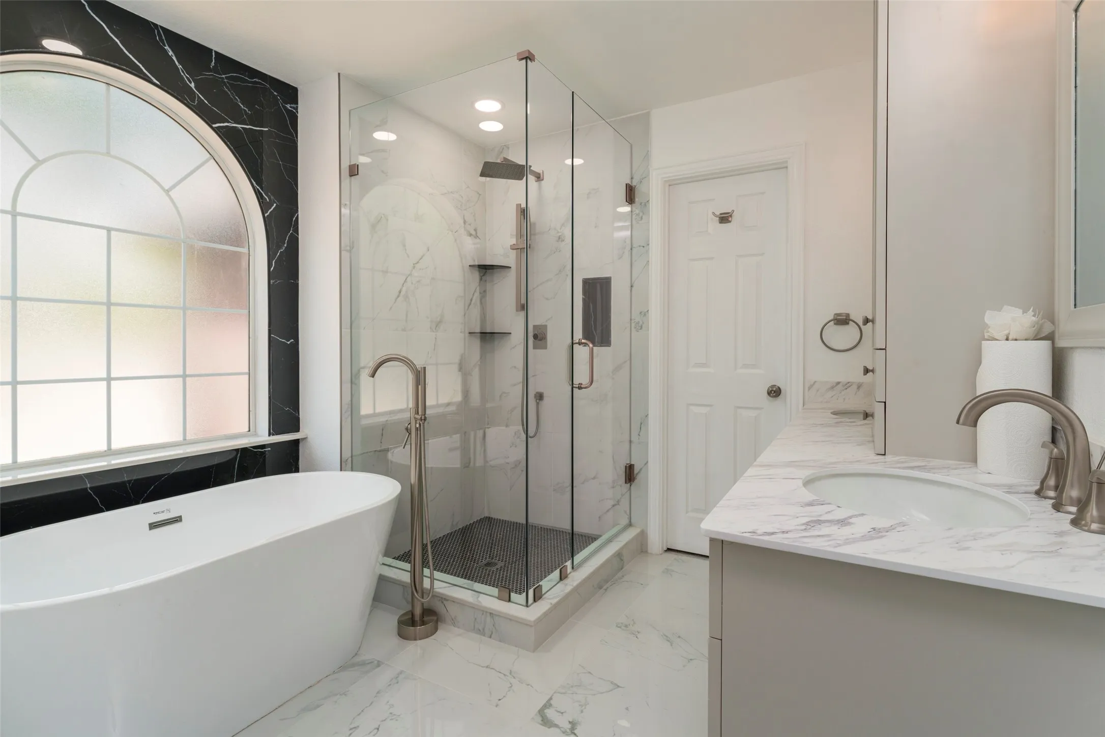Freestanding soaking tub with floor-mounted faucet, arched window, glass-enclosed shower, dual vanity with undermount sink, and marble-patterned flooring