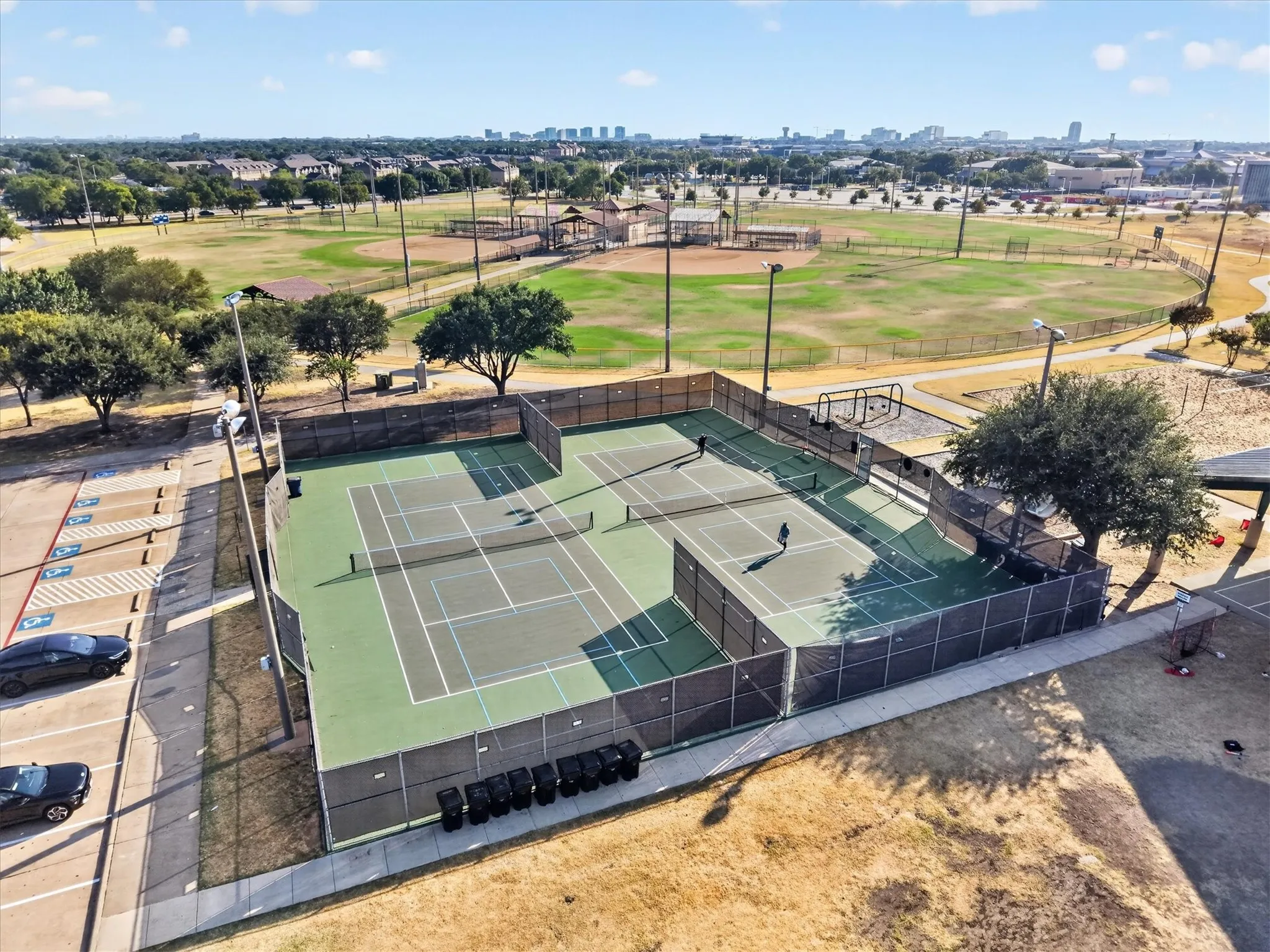 Fenced tennis courts with hard surfacing, adjacent to a large open field and a paved parking area