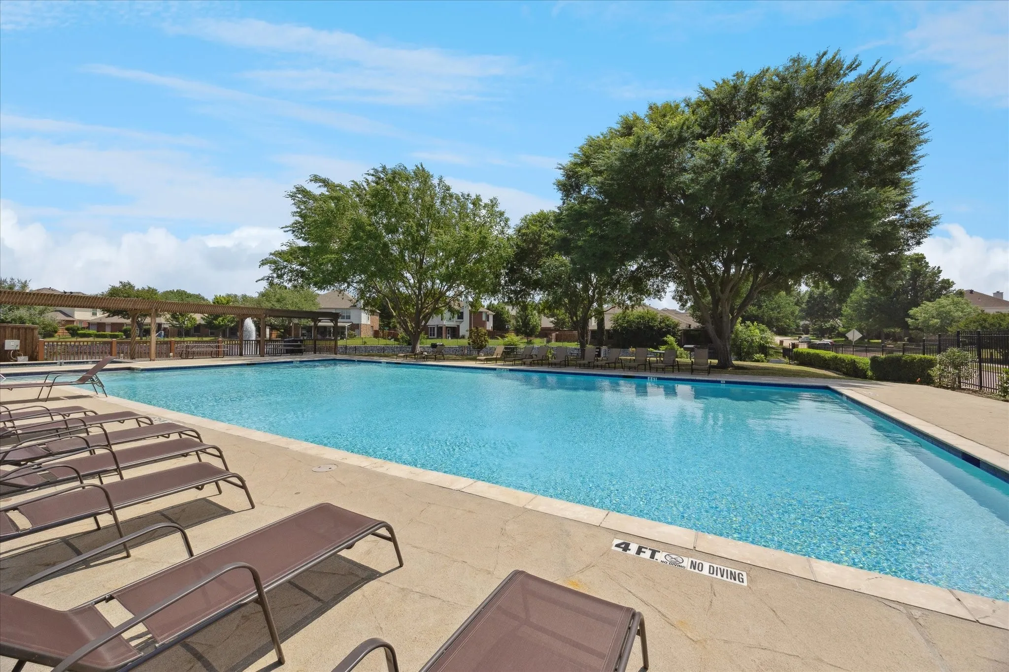 Expansive swimming pool with concrete decking, surrounding trees, and a distant pergola structure