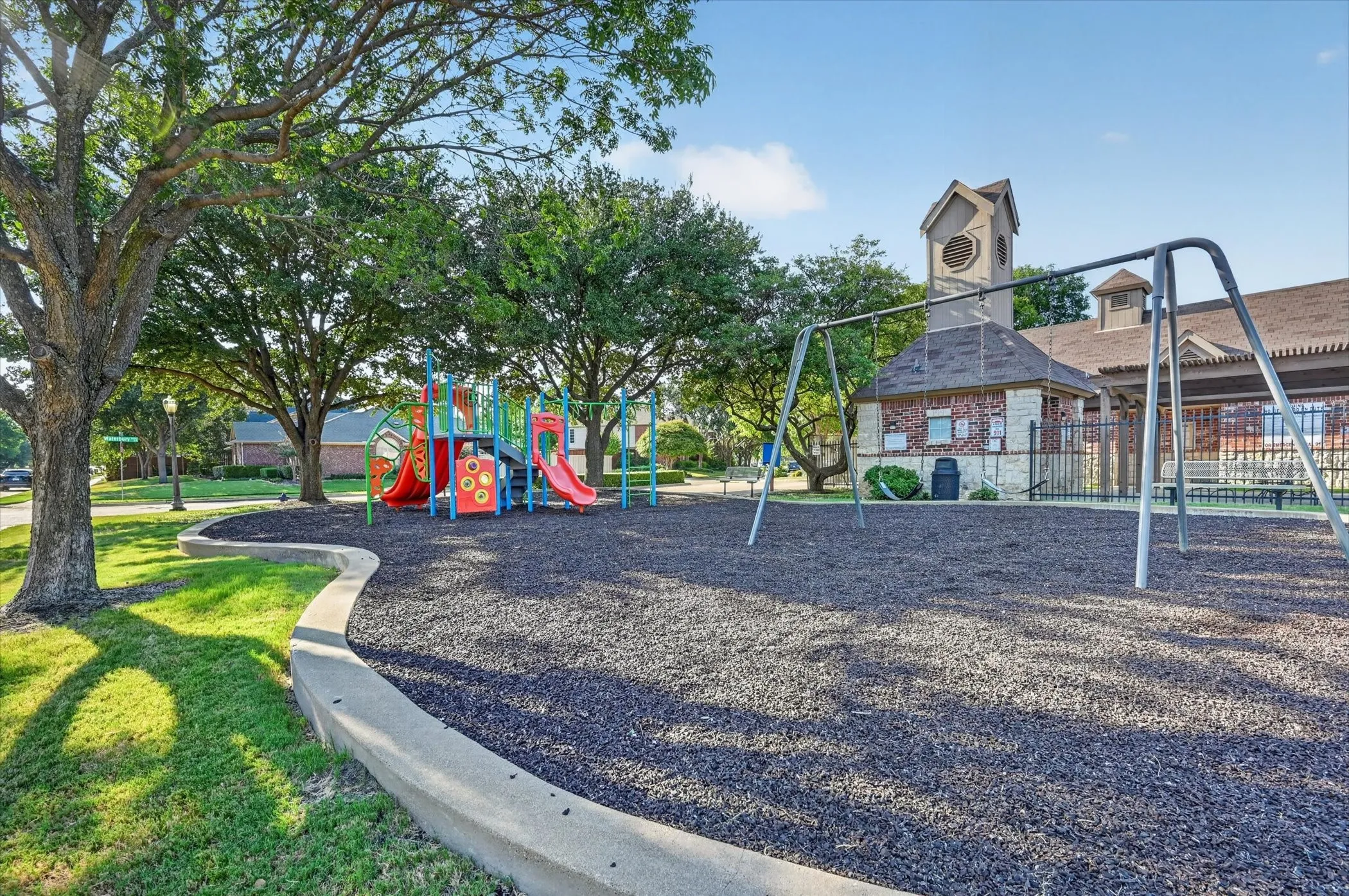 Recreational area featuring a playground with slides, swing sets, mature trees, a manicured lawn, and a community building with a clock tower