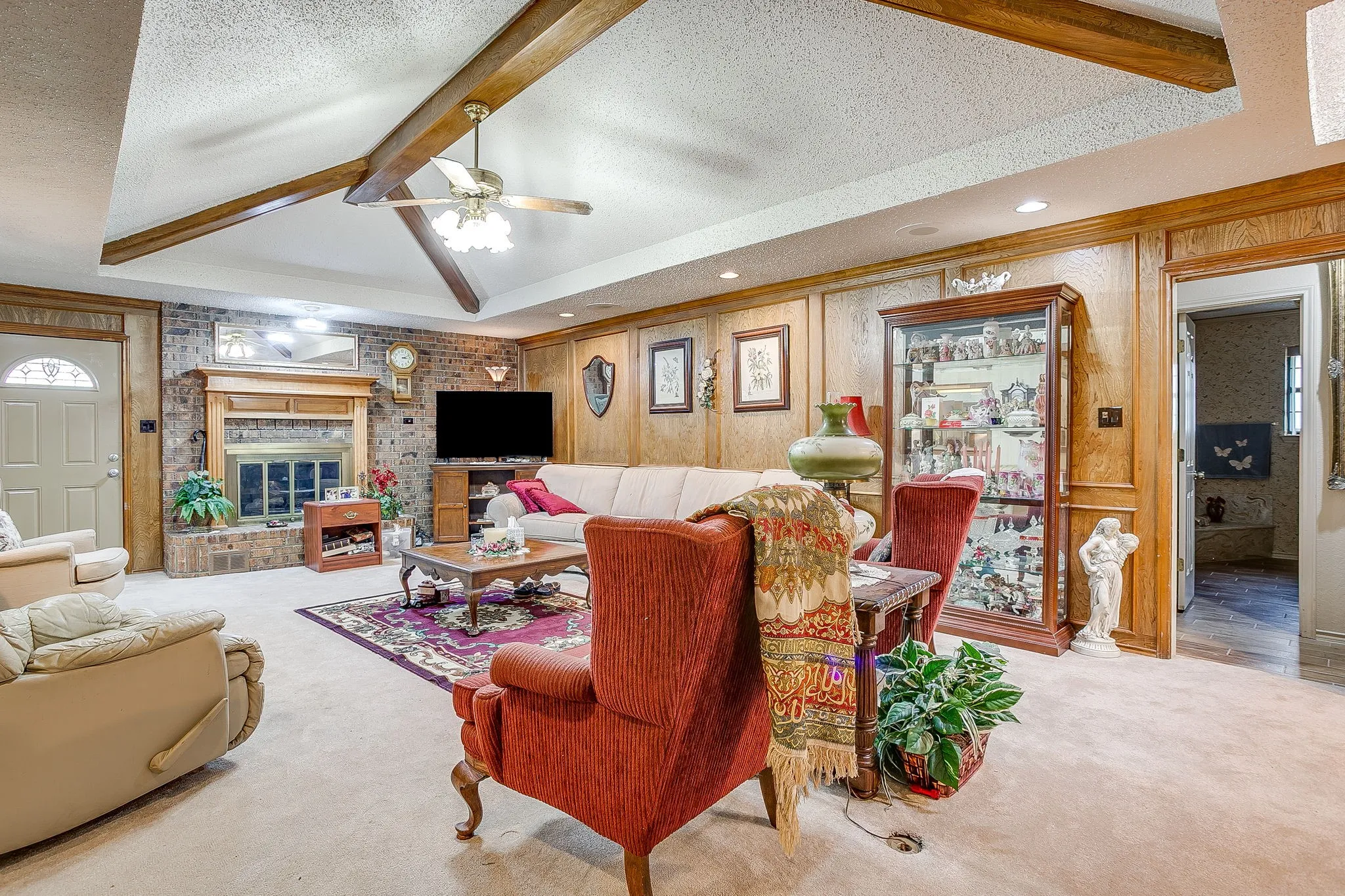 Recessed ceiling with exposed wood beams, brick-surround fireplace with wood mantel, wood paneling, and a ceiling fan