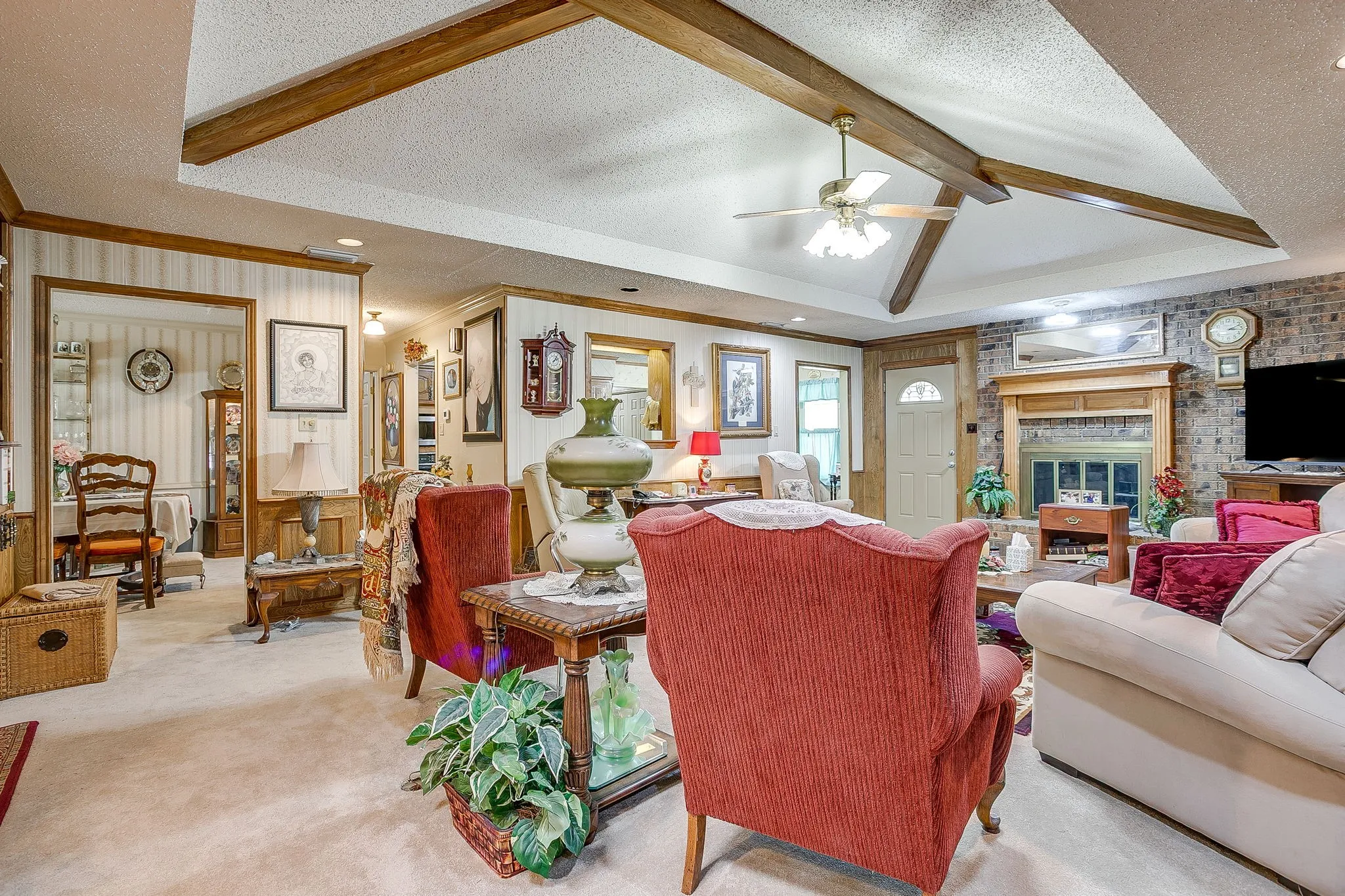 Vaulted ceiling with exposed wood beams, ceiling fan, brick-faced fireplace with wood mantel, and wood-trim details
