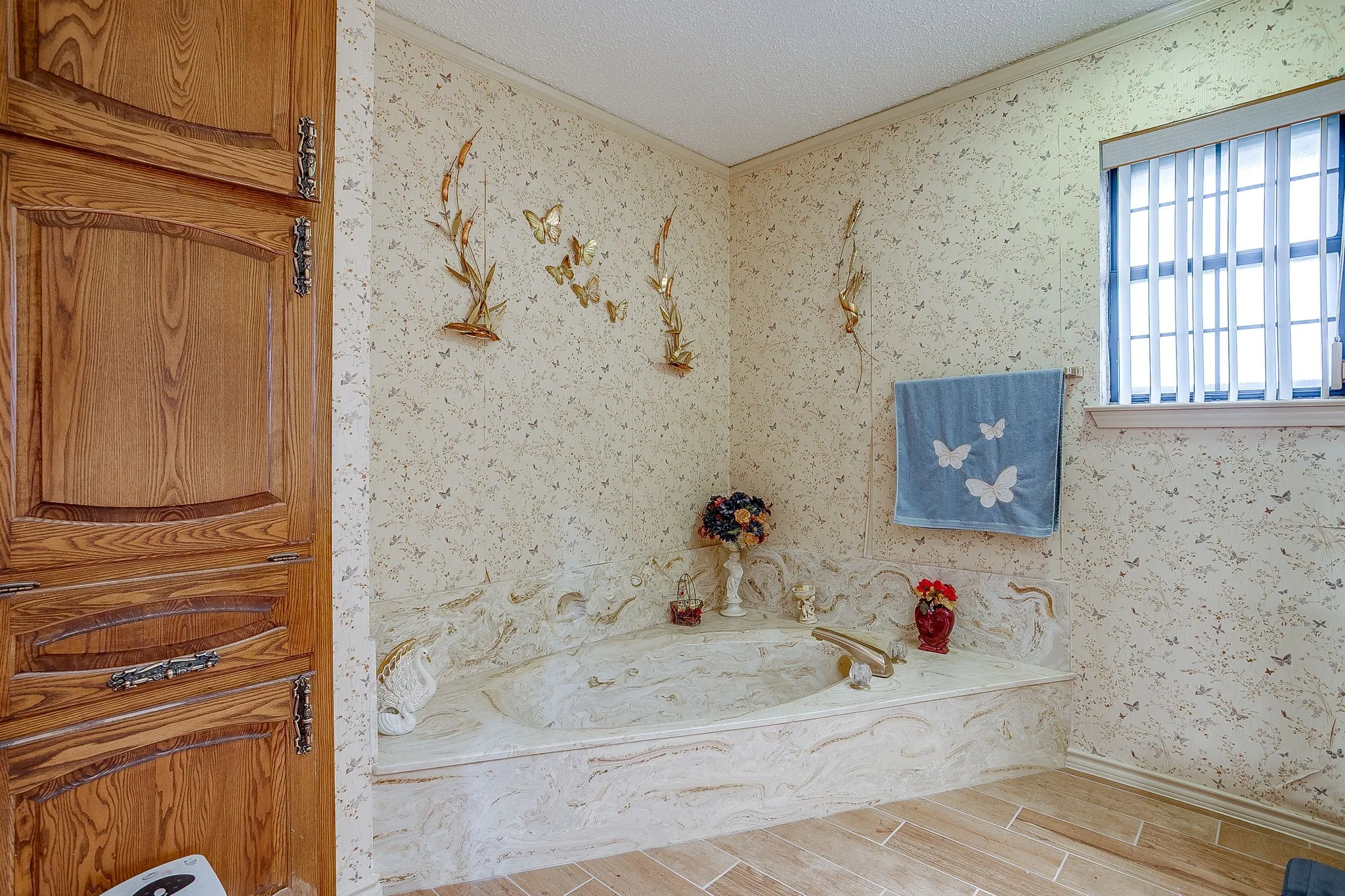Built-in soaking tub with decorative surround, featuring a window with horizontal blinds, wood-finish flooring, and a large wooden storage cabinet