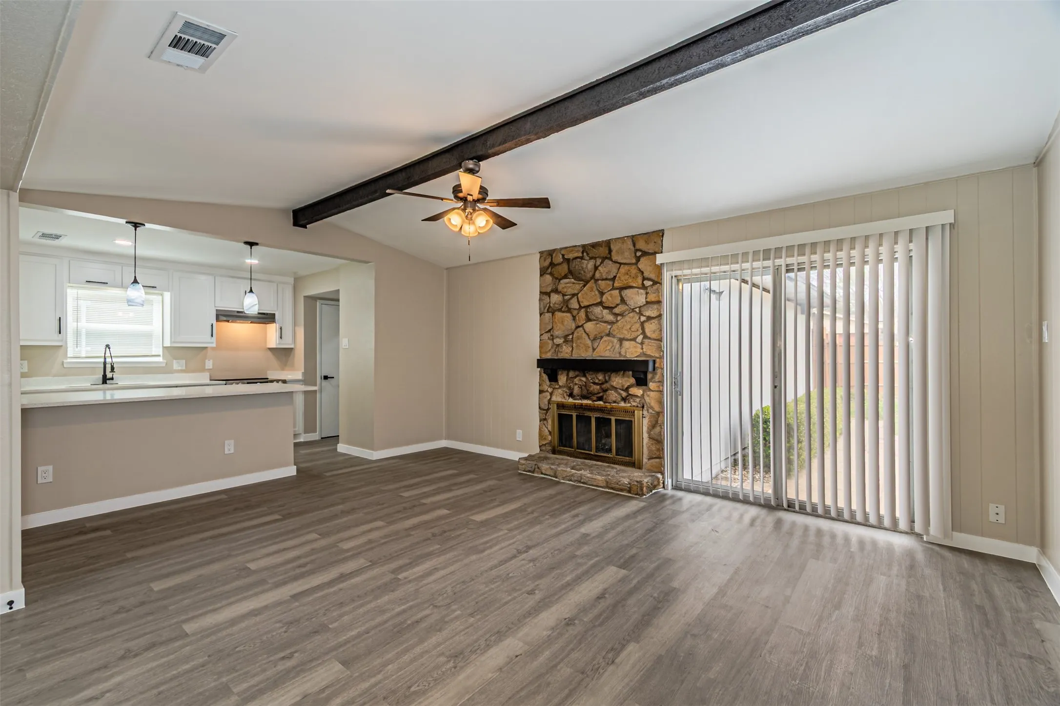 Stone-clad fireplace with mantel, sliding glass door with vertical blinds, wood-finish flooring, vaulted ceiling with exposed beam, and a kitchen pass-through with pendant lighting