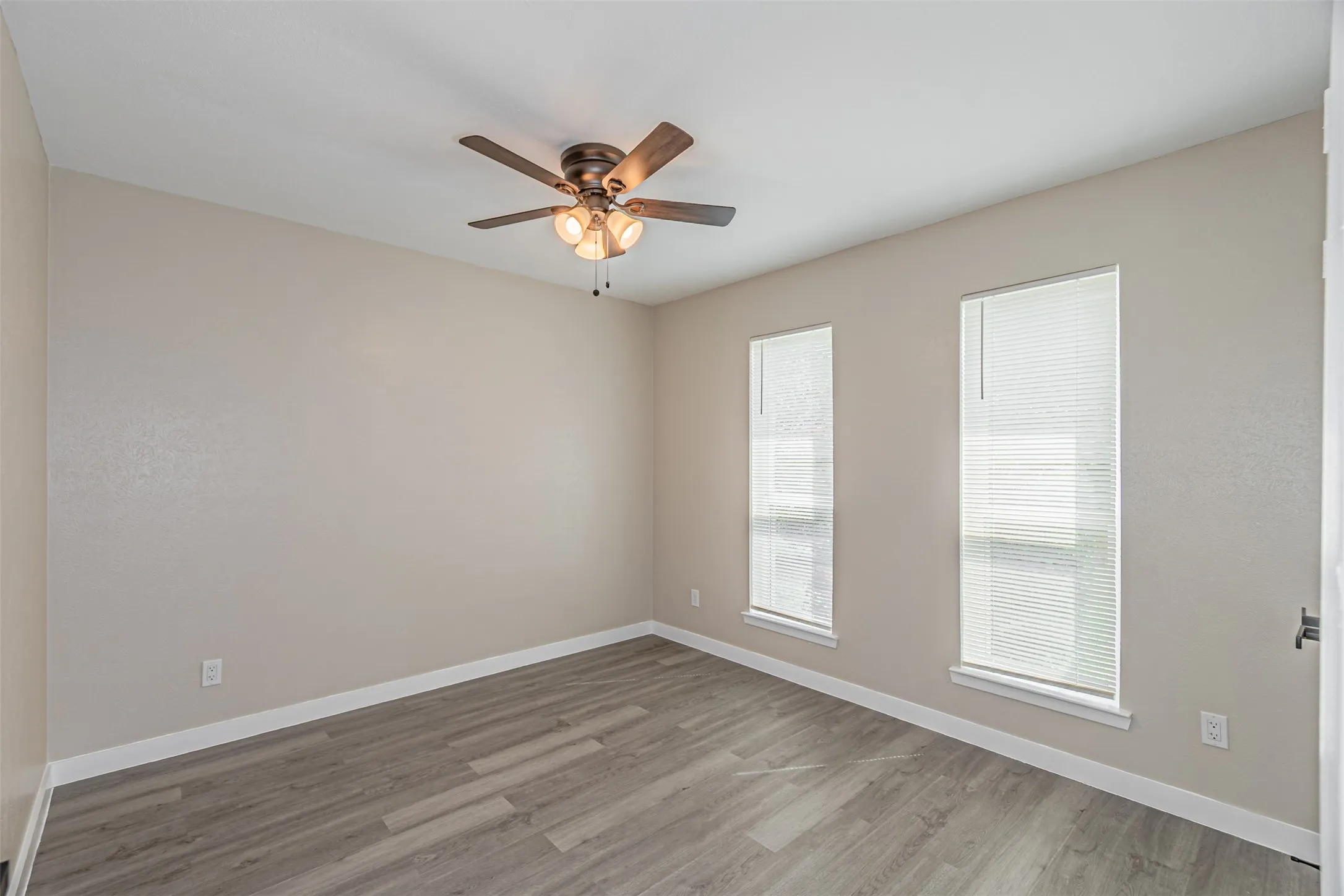 Neutral wall finishes, wood-finish flooring, two large windows, white window blinds, and a ceiling fan with integrated lighting