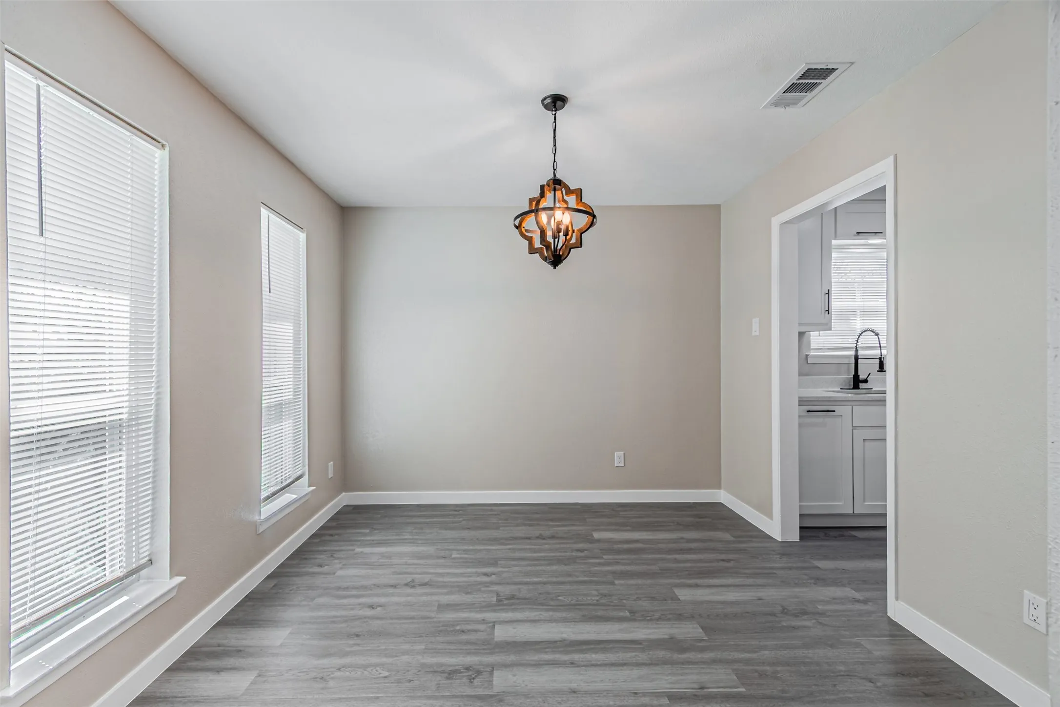 Wood-finish flooring, two windows with blinds, a decorative ceiling fixture, and a doorway revealing white cabinetry and a dark faucet