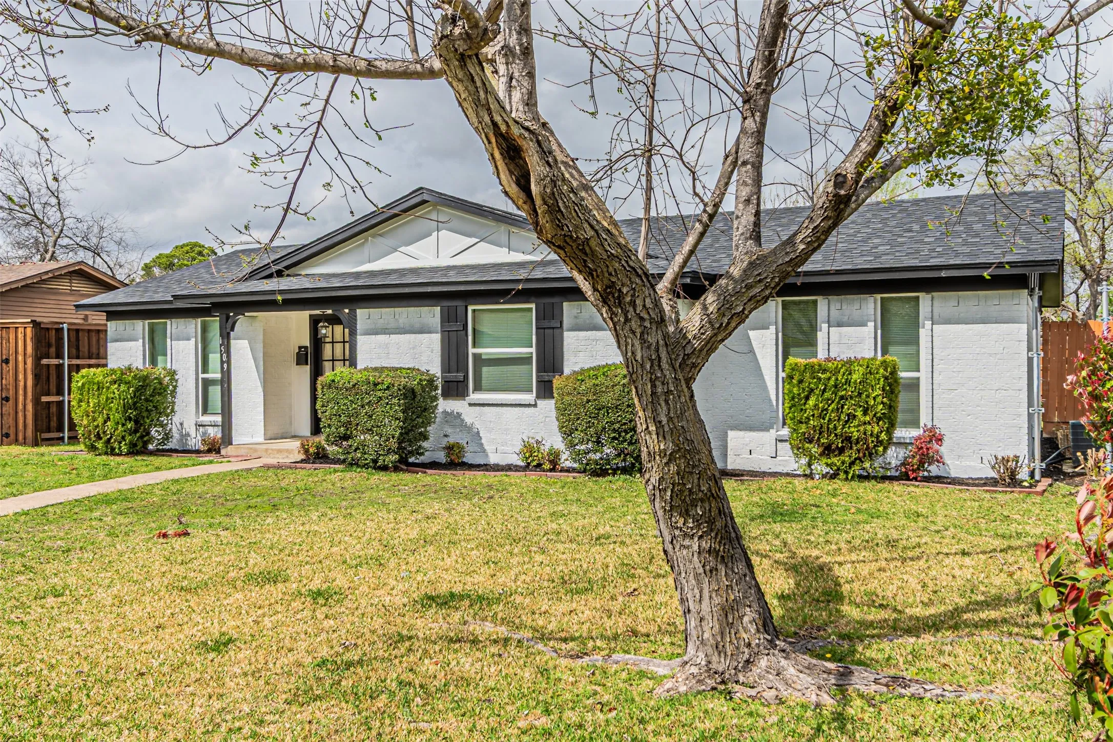 White painted brick exterior with dark shutters, covered front entry, established front lawn, and mature landscaping
