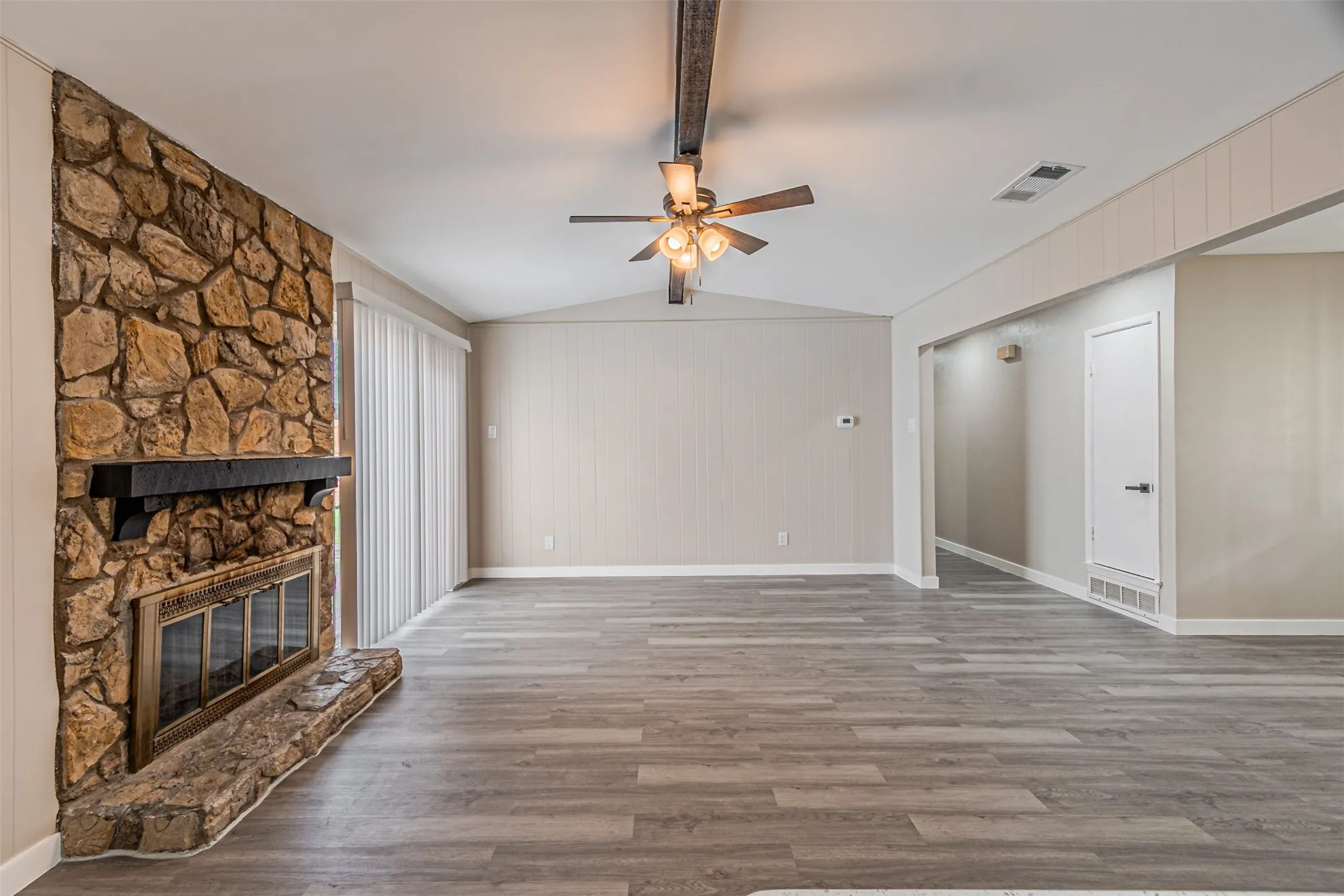 Spacious living area featuring a stone fireplace with hearth, wood-finish flooring, vaulted ceiling with exposed beam, and a ceiling fan