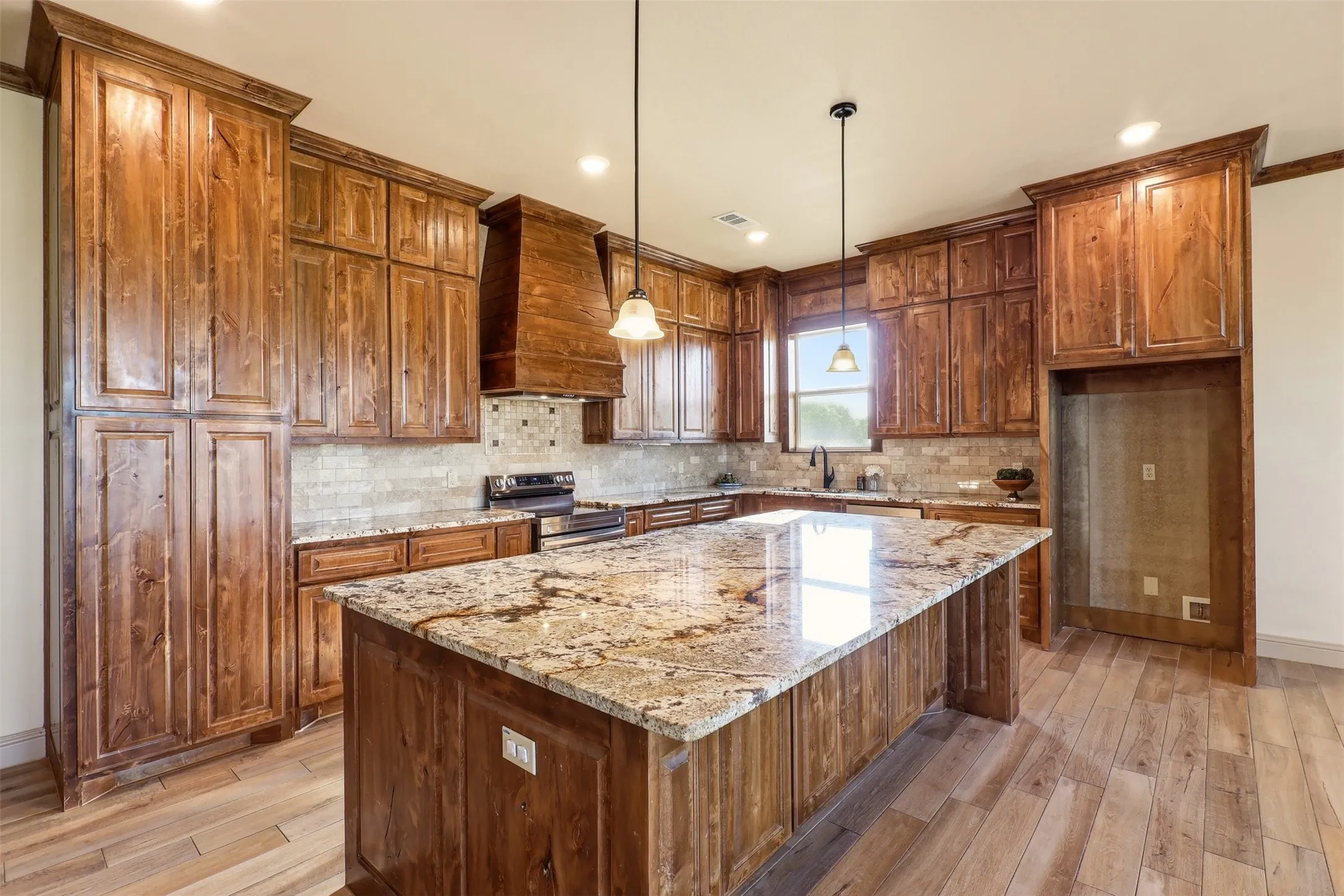 Expansive kitchen island with a stone countertop, extensive wood cabinetry, wood-finish flooring, built-in range, and a tiled backsplash