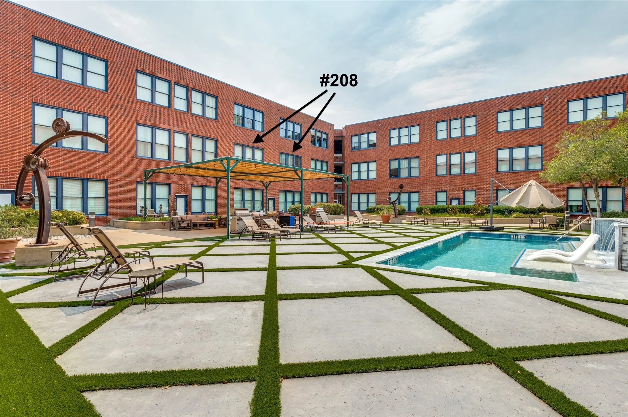 Modern residential courtyard featuring a resort-style heated swimming pool, expansive concrete hardscape with integrated turf sections, lounge seating areas, and a contemporary red-brick building facade with multi-story fenestration and manicured landscaping