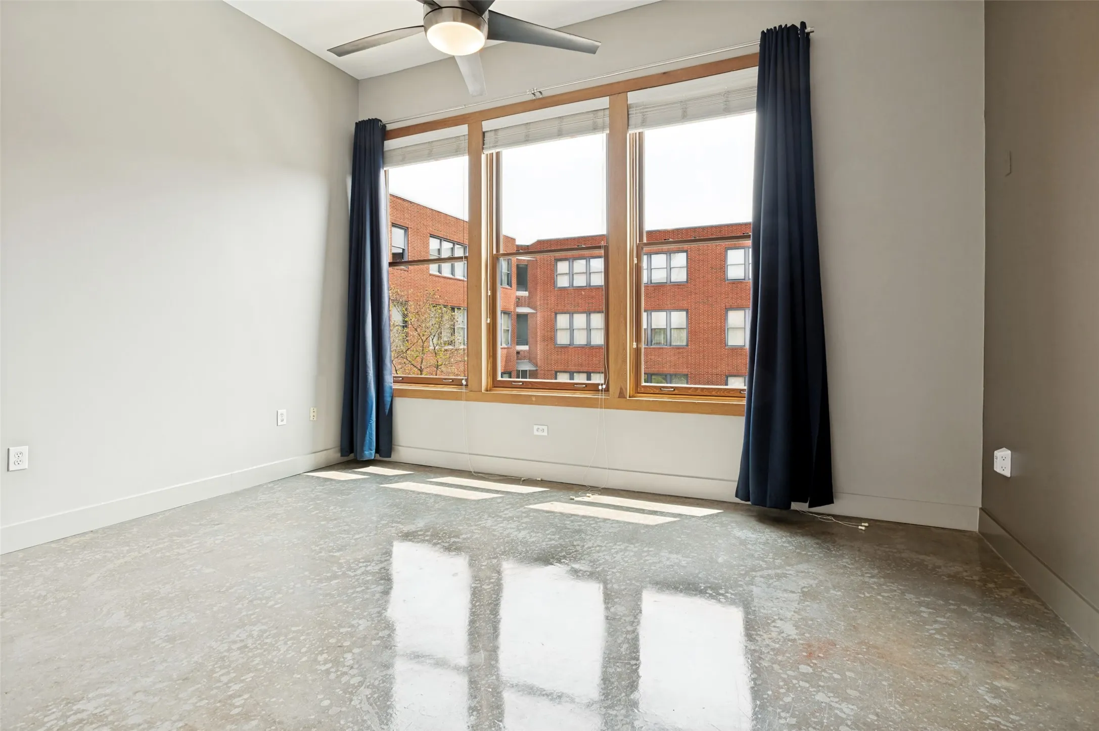 Polished concrete flooring, large window with wood-finish framing, ceiling fan with integrated lighting, and neutral wall tones