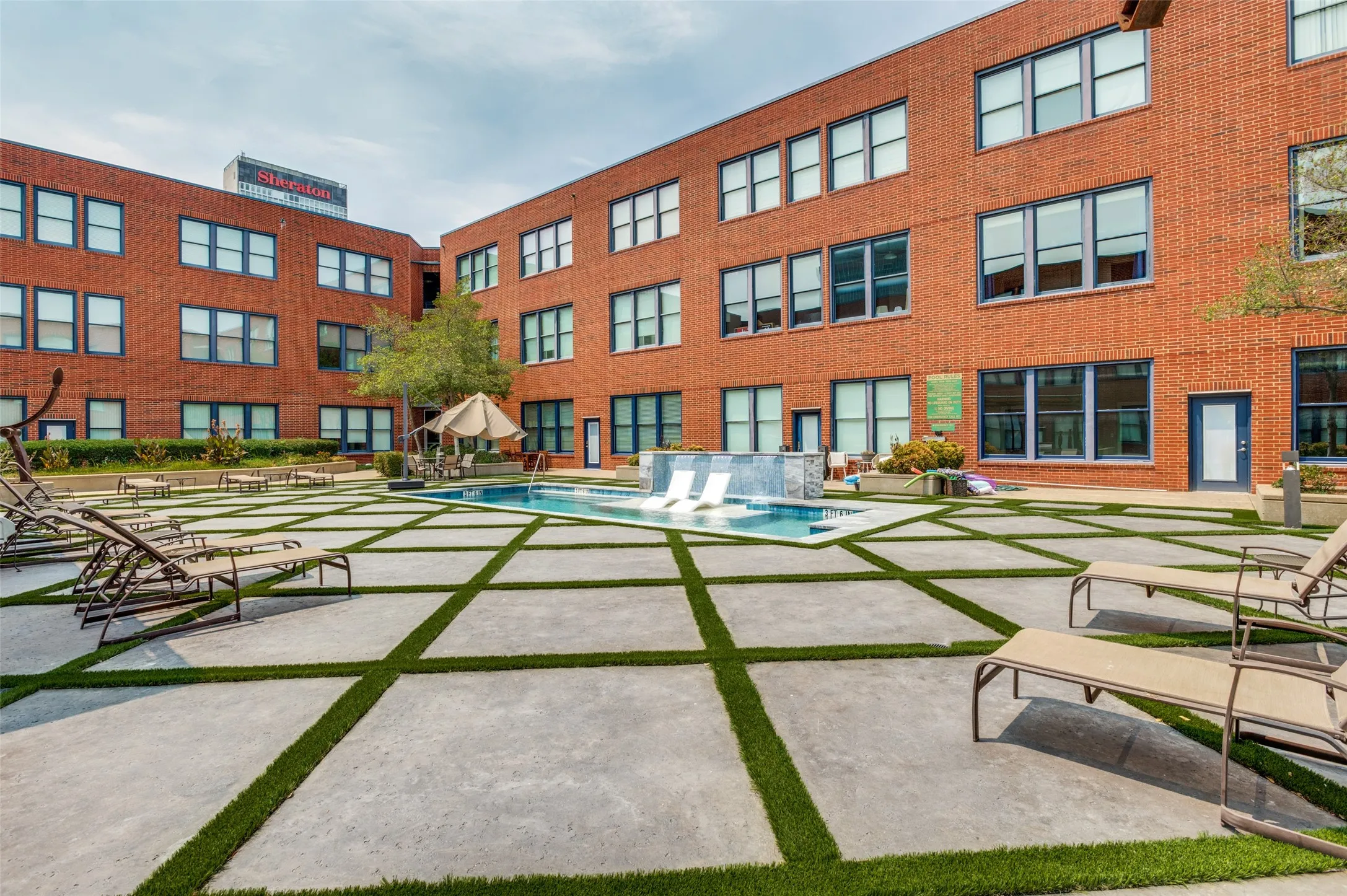 Brick exterior building with multiple windows, central swimming pool, integrated water feature, and a concrete paver patio with artificial turf accents