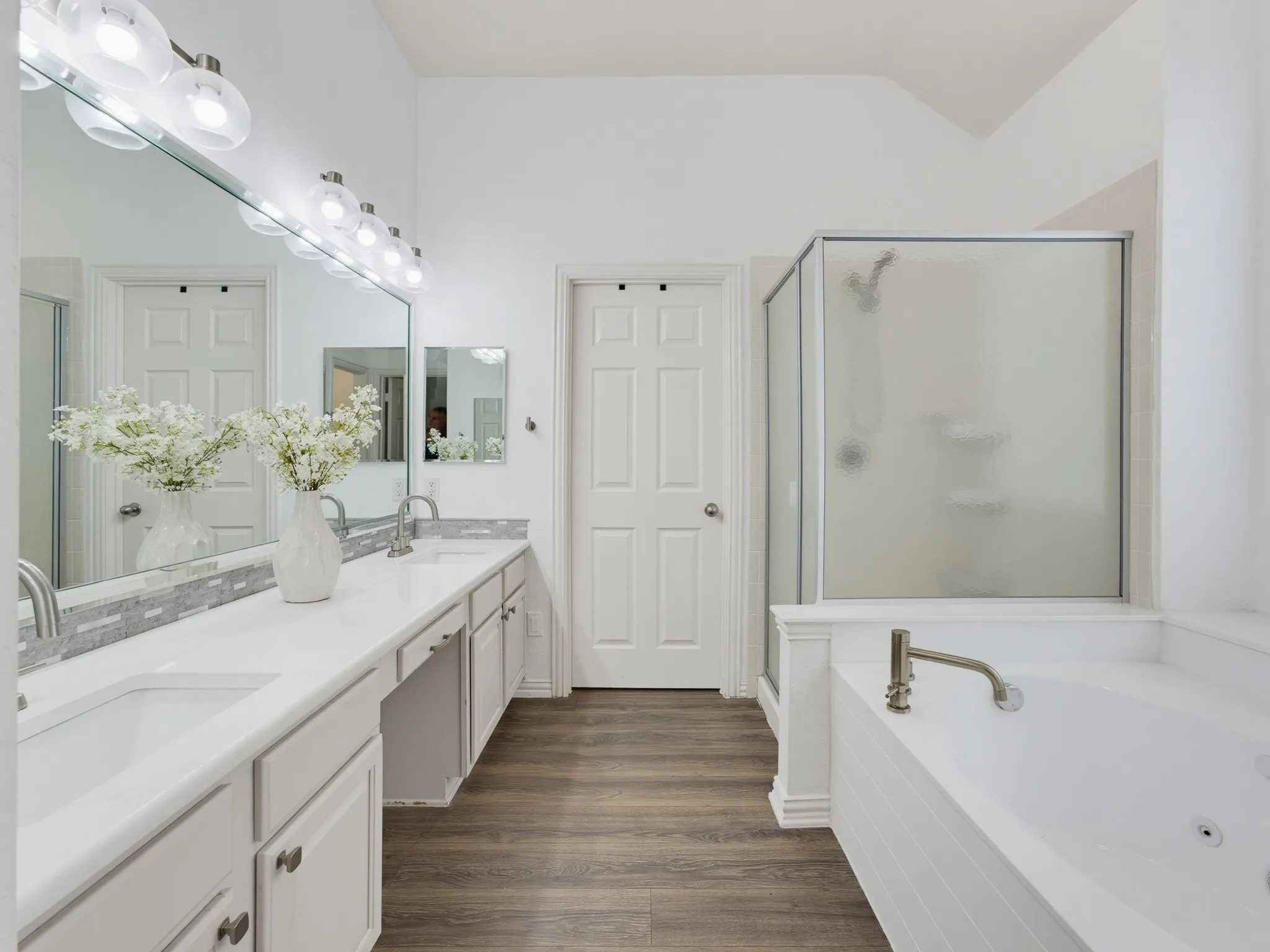 Dual vanity with integrated sinks, full-width mirror, separate shower enclosure, soaking tub, and wood-finish flooring