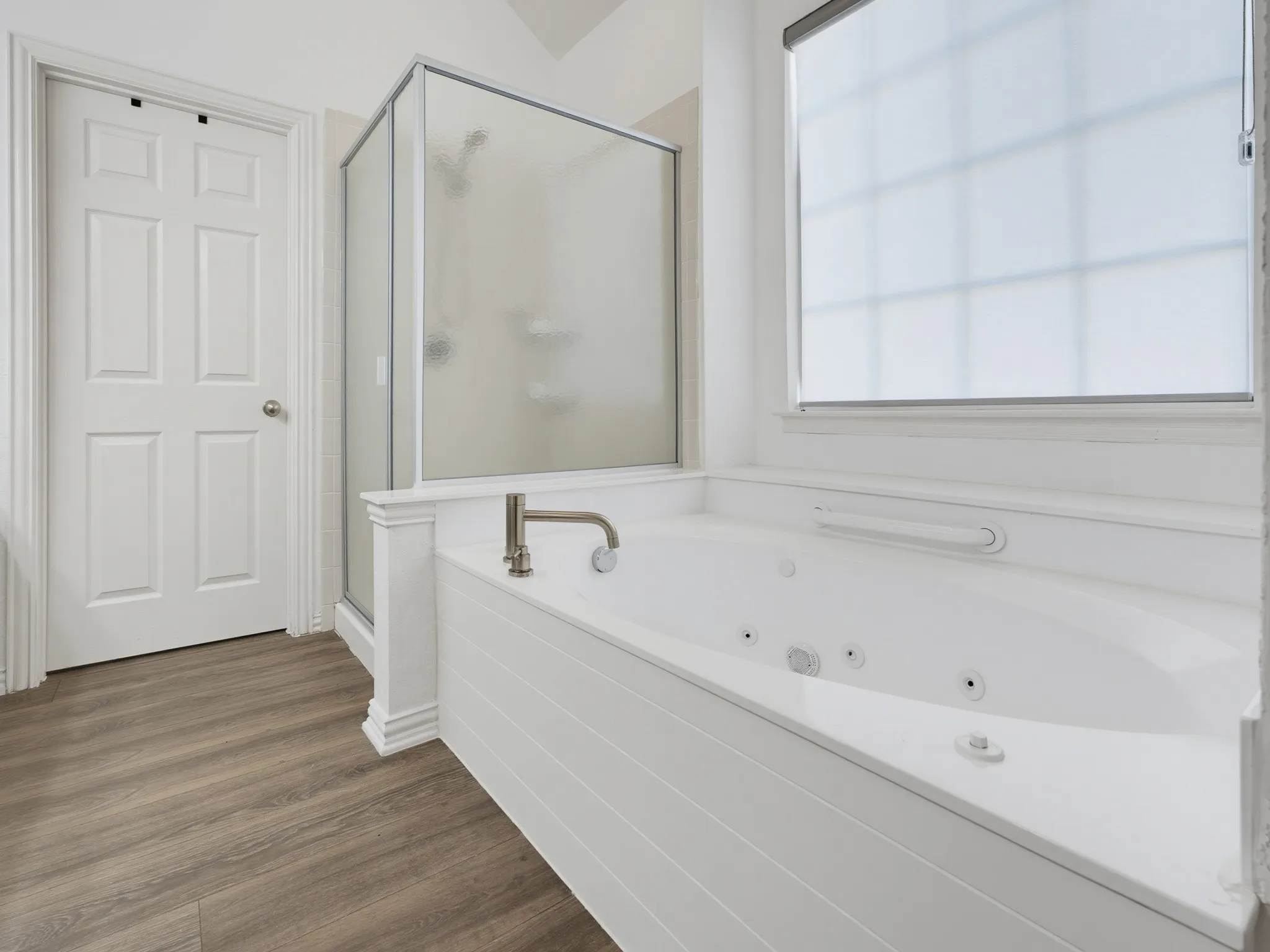 Soaking tub with brushed nickel fixtures, glass-enclosed shower, wood-finish flooring, and a privacy window treatment