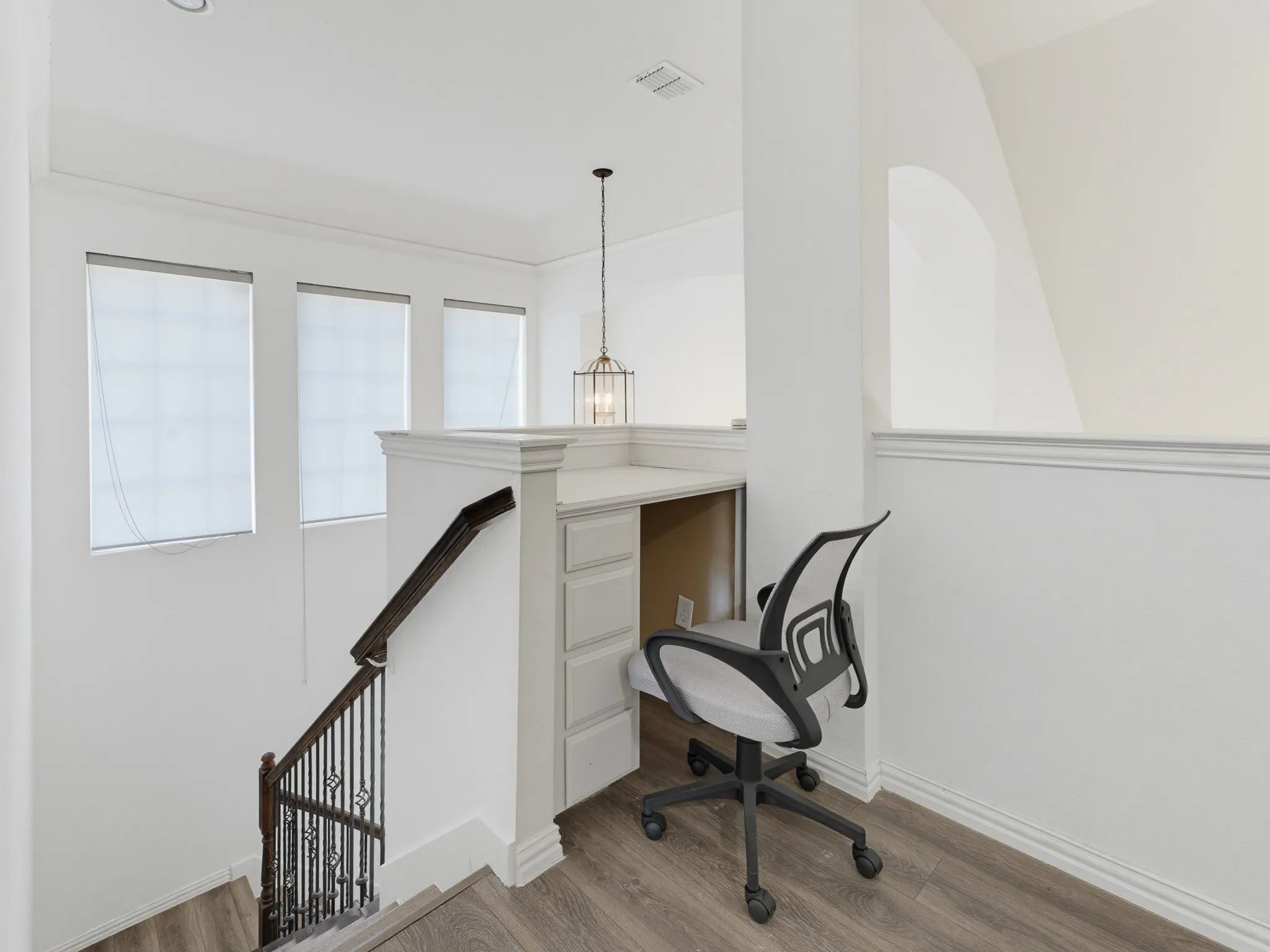 Built-in desk with paneled drawers, wood-finish flooring, staircase with dark wood handrail and wrought iron balusters, and three windows with roller shades