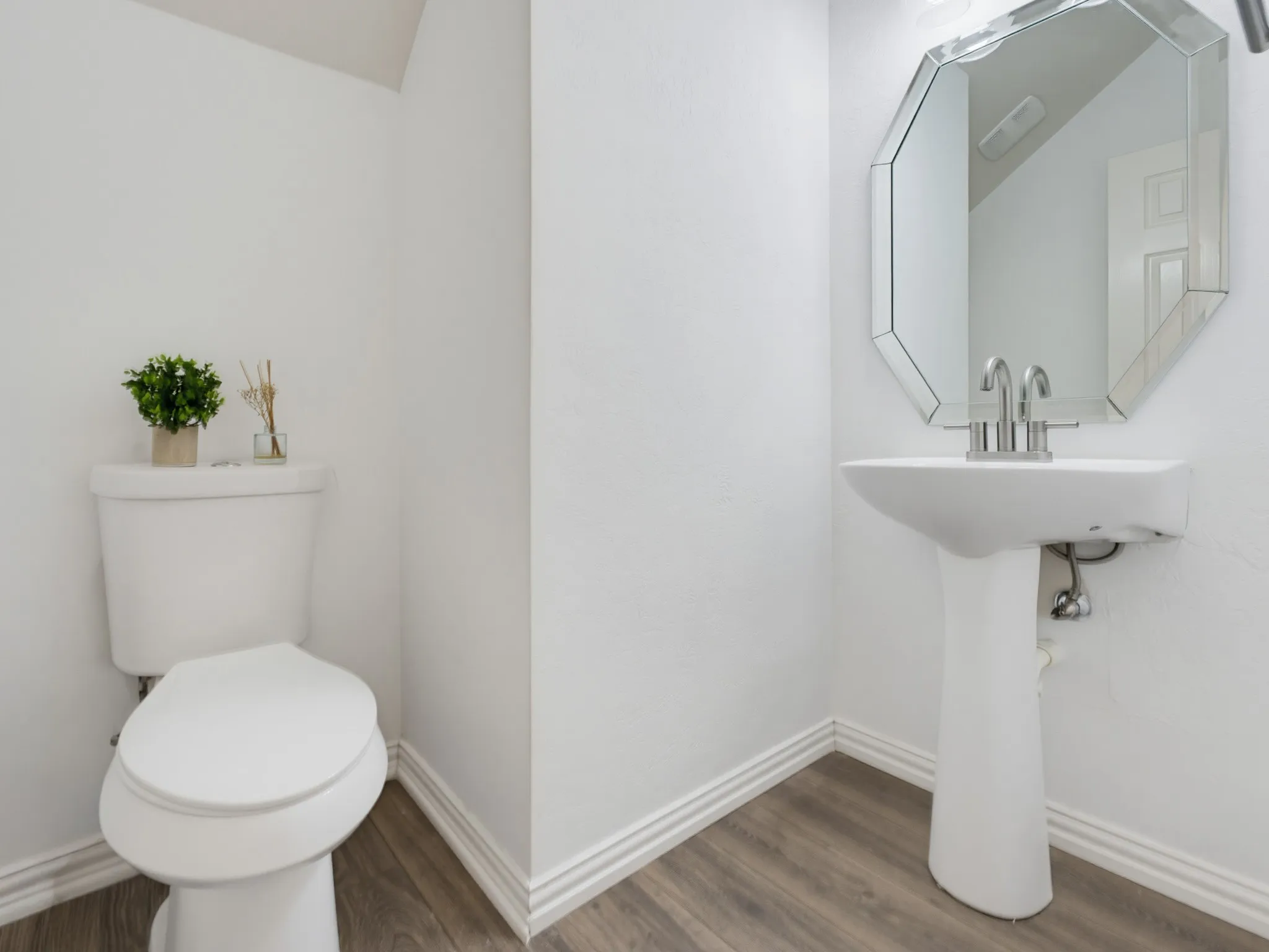 Pedestal sink with a polished chrome faucet, octagonal frameless mirror, and wood-finish flooring