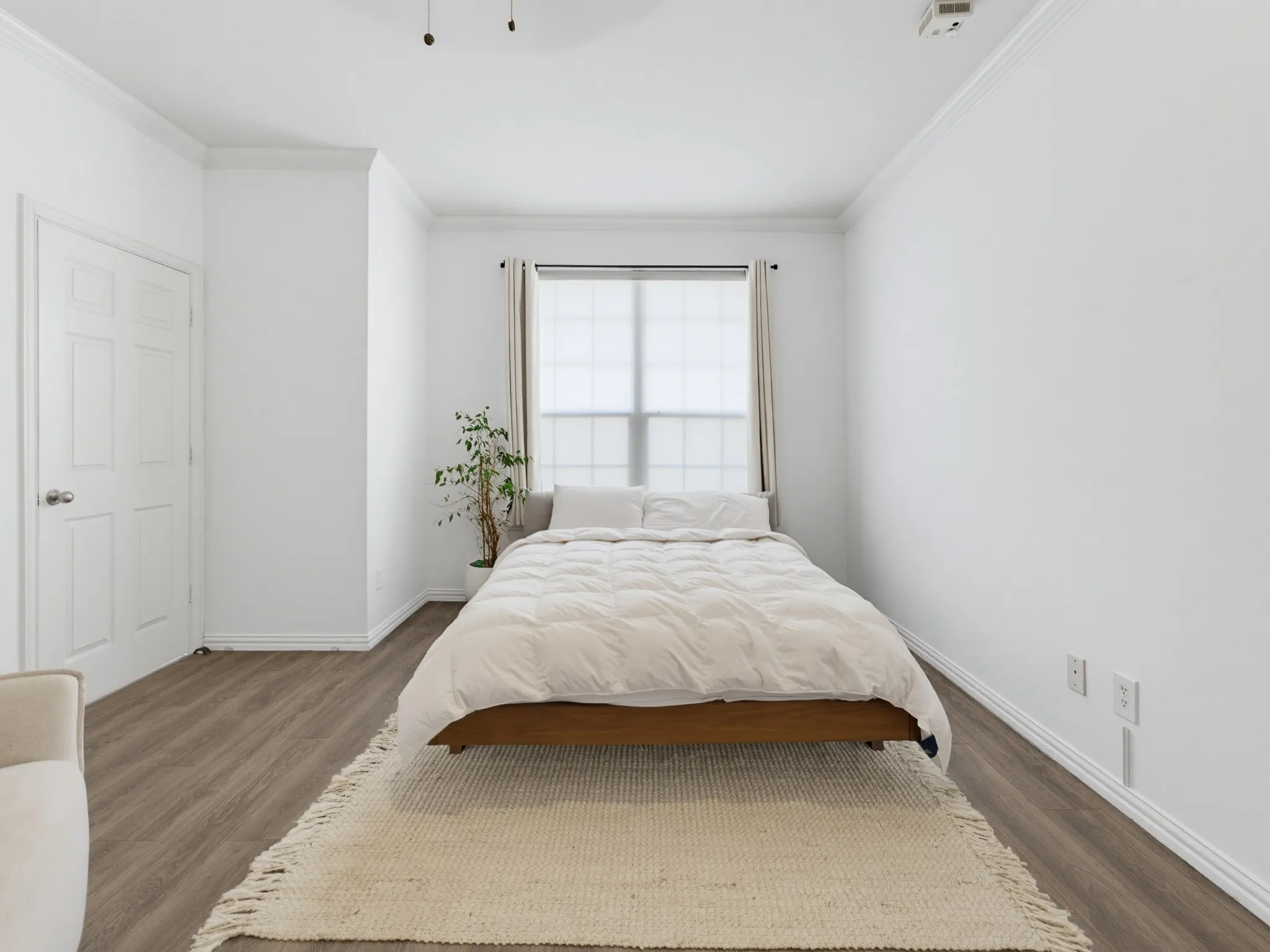 Wood-finish flooring, white paneled door, crown molding, window with roller shade, and white walls