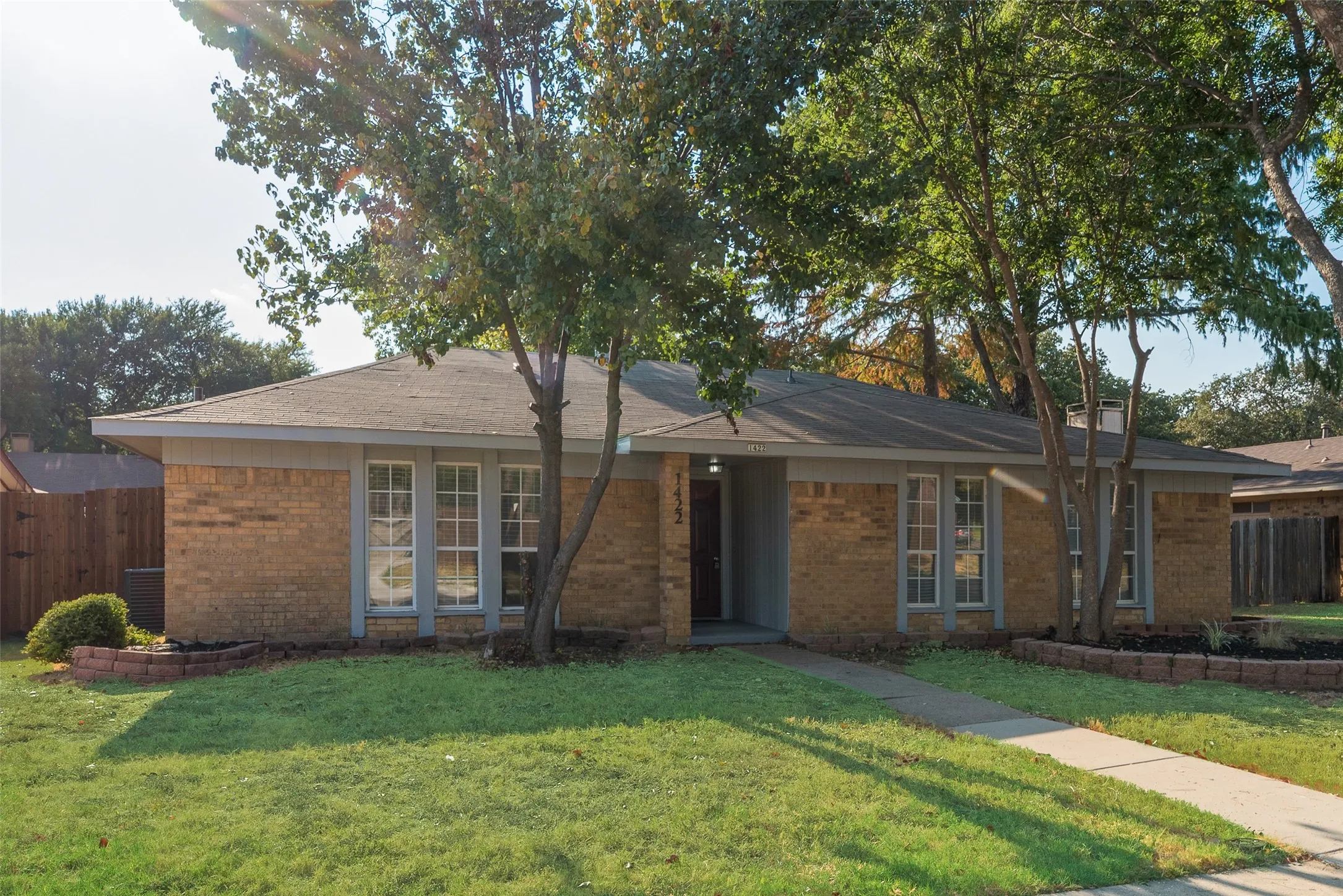 Brick exterior facade with a dark shingle roof, multiple mature trees, and a concrete pathway leading to the front entry