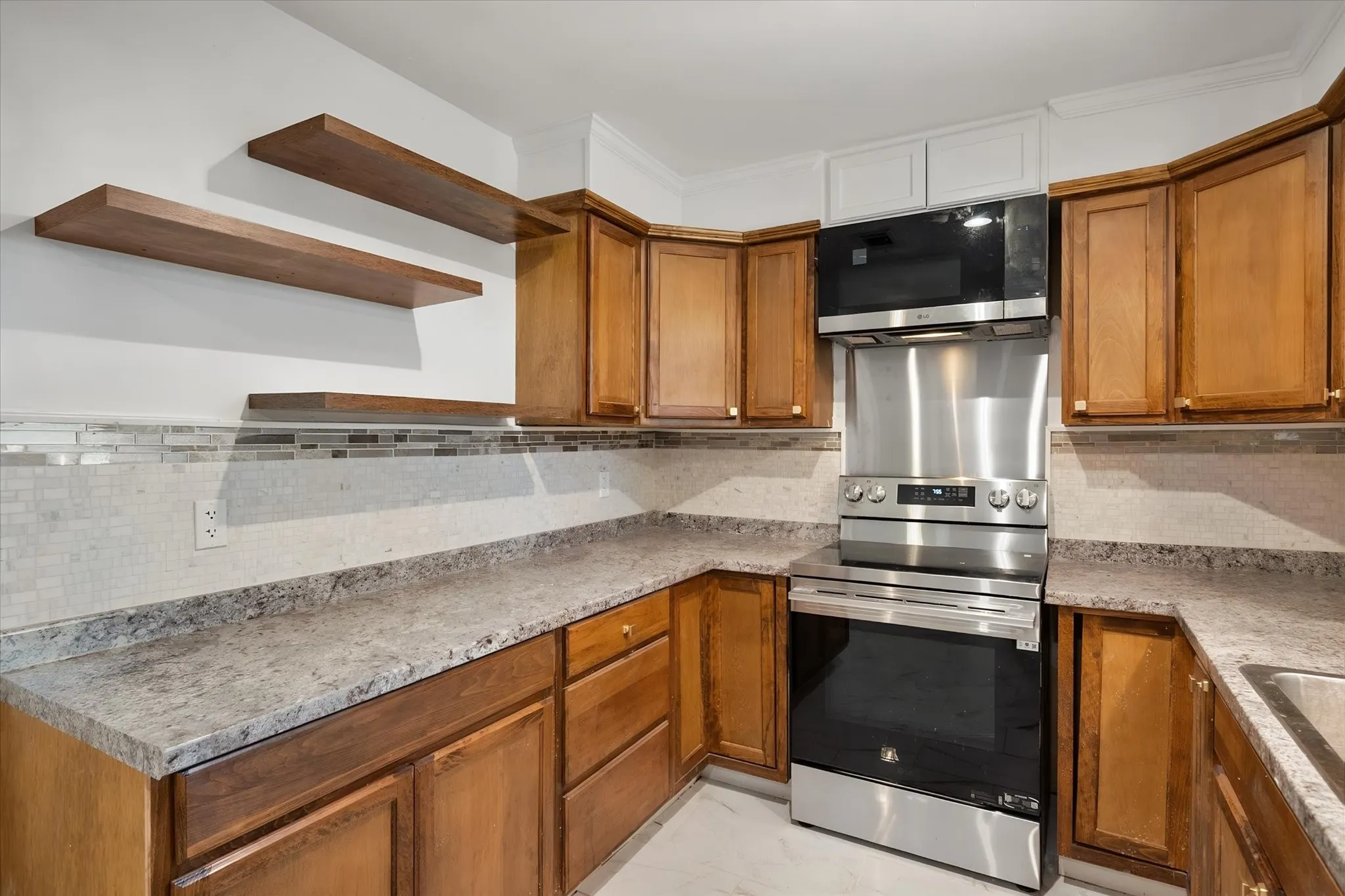 Kitchen featuring warm wood cabinetry, granite-style countertops, mosaic tile backsplash, stainless steel appliances, and floating wood shelves