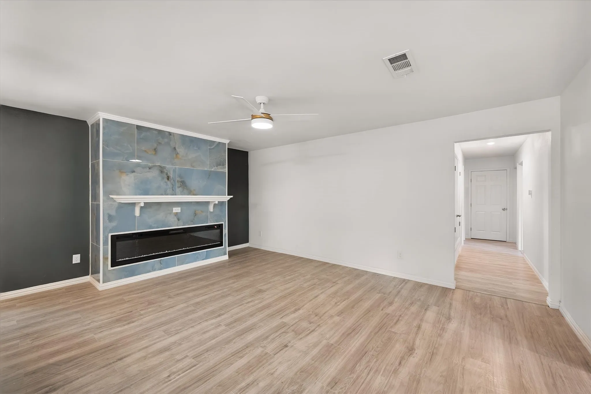 Wood-finish flooring, accent wall with a tiled electric fireplace, decorative mantel, and ceiling fan