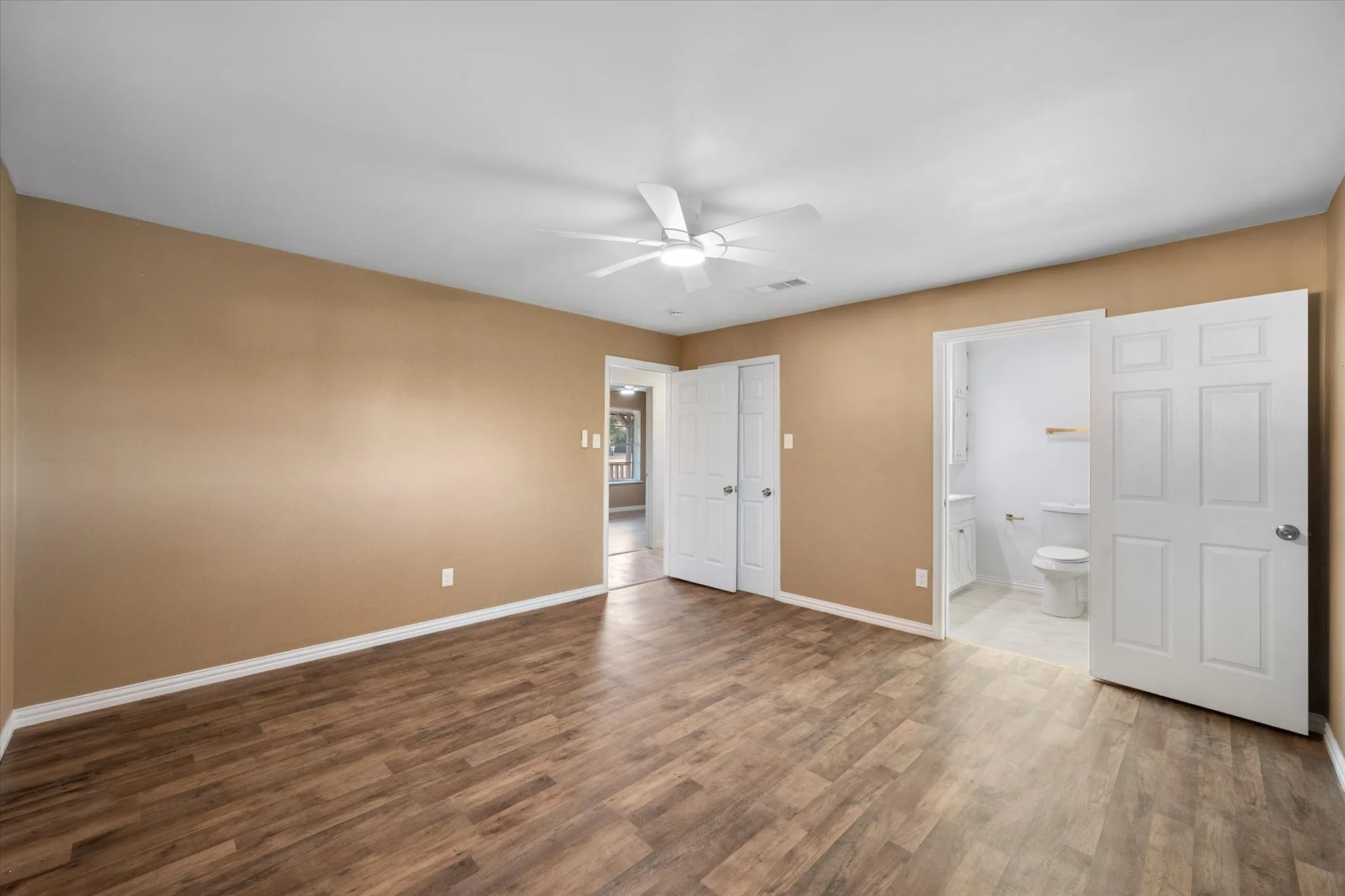Wood-finish flooring, ceiling fan, white paneled doors, and an attached bathroom with a toilet and vanity