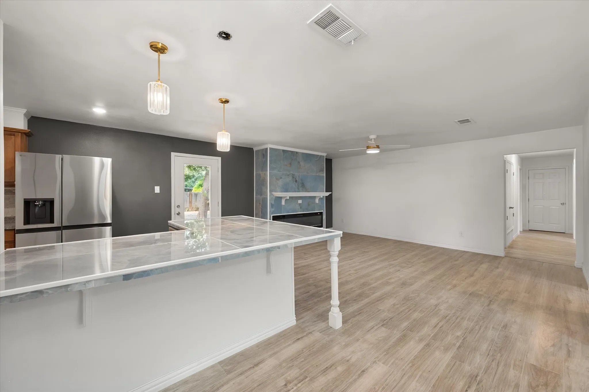 Open-concept space featuring wood-finish flooring, a white kitchen island with a stone-finish countertop, stainless steel refrigerator, and a fireplace with a stone-finish surround