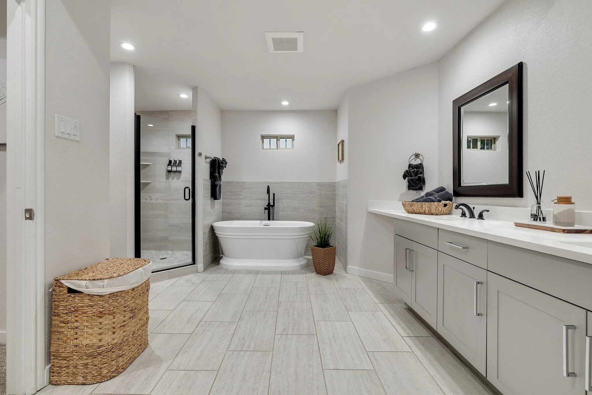 Freestanding soaking tub, large vanity with light gray cabinetry, wall-mounted mirror, and a glass-enclosed shower with black trim