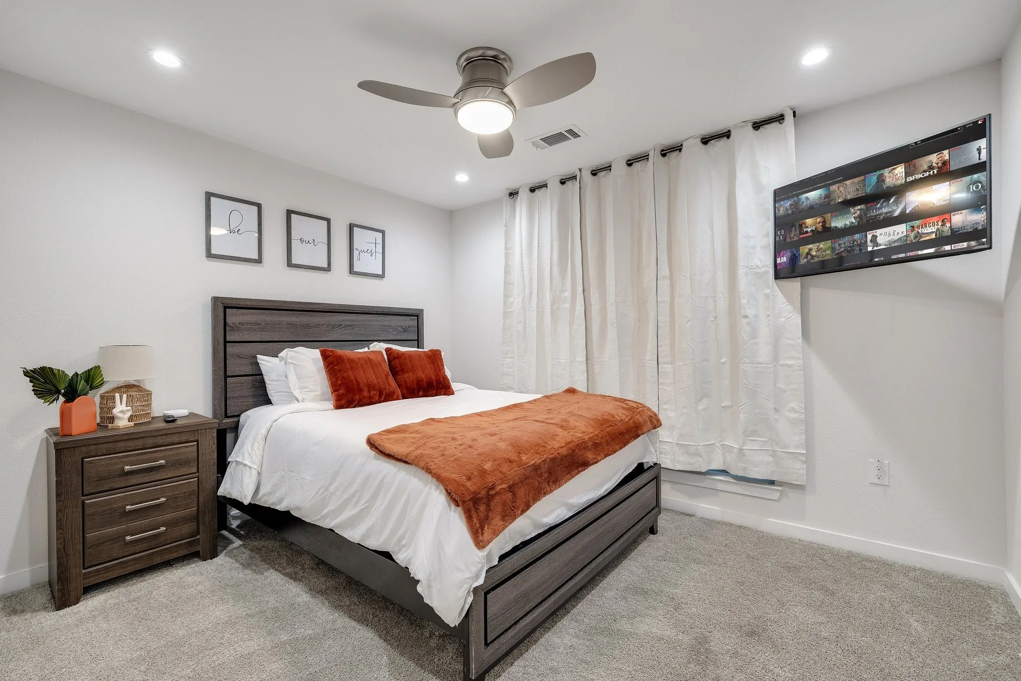 Master bedroom featuring a wood-finish platform bed with upholstered headboard, soft white bedding with decorative throw pillows, coordinating wood-finish nightstand with drawer storage, recessed ceiling lighting, ceiling fan with integrated light fixture, wall-mounted flat-screen television, floor-to-ceiling sheer curtains, and neutral carpet flooring throughout
