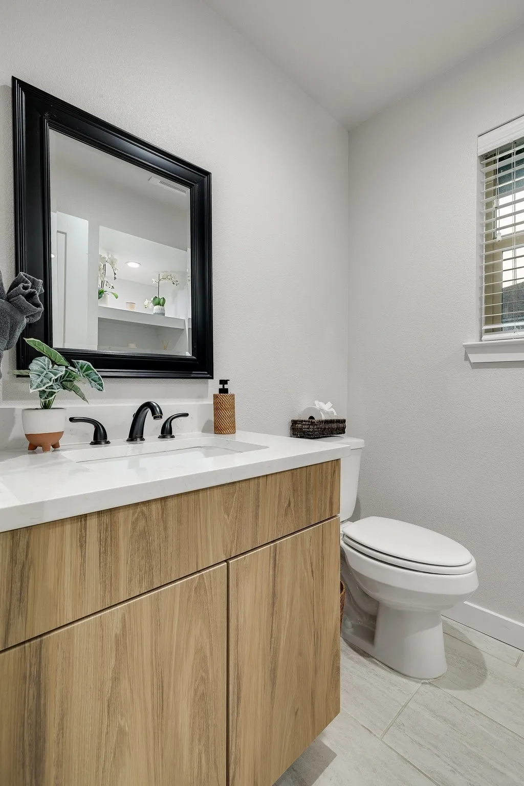 Wood-finish vanity with a white countertop and undermount sink, accompanied by a framed mirror, toilet, and a window with blinds