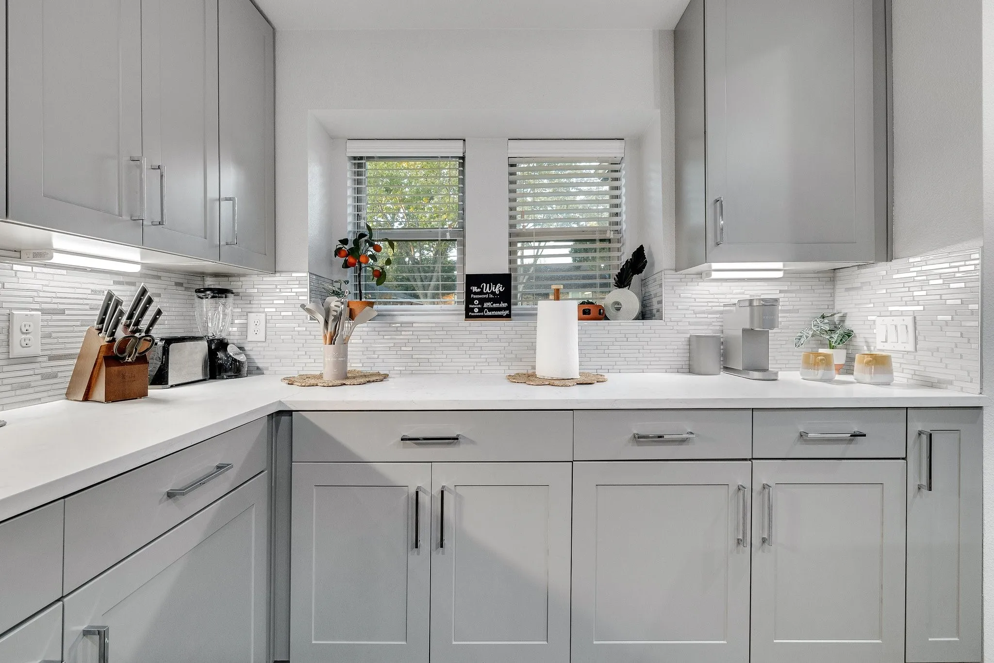 White countertops, light gray cabinetry, under-cabinet lighting, mosaic tile backsplash, and stainless steel hardware