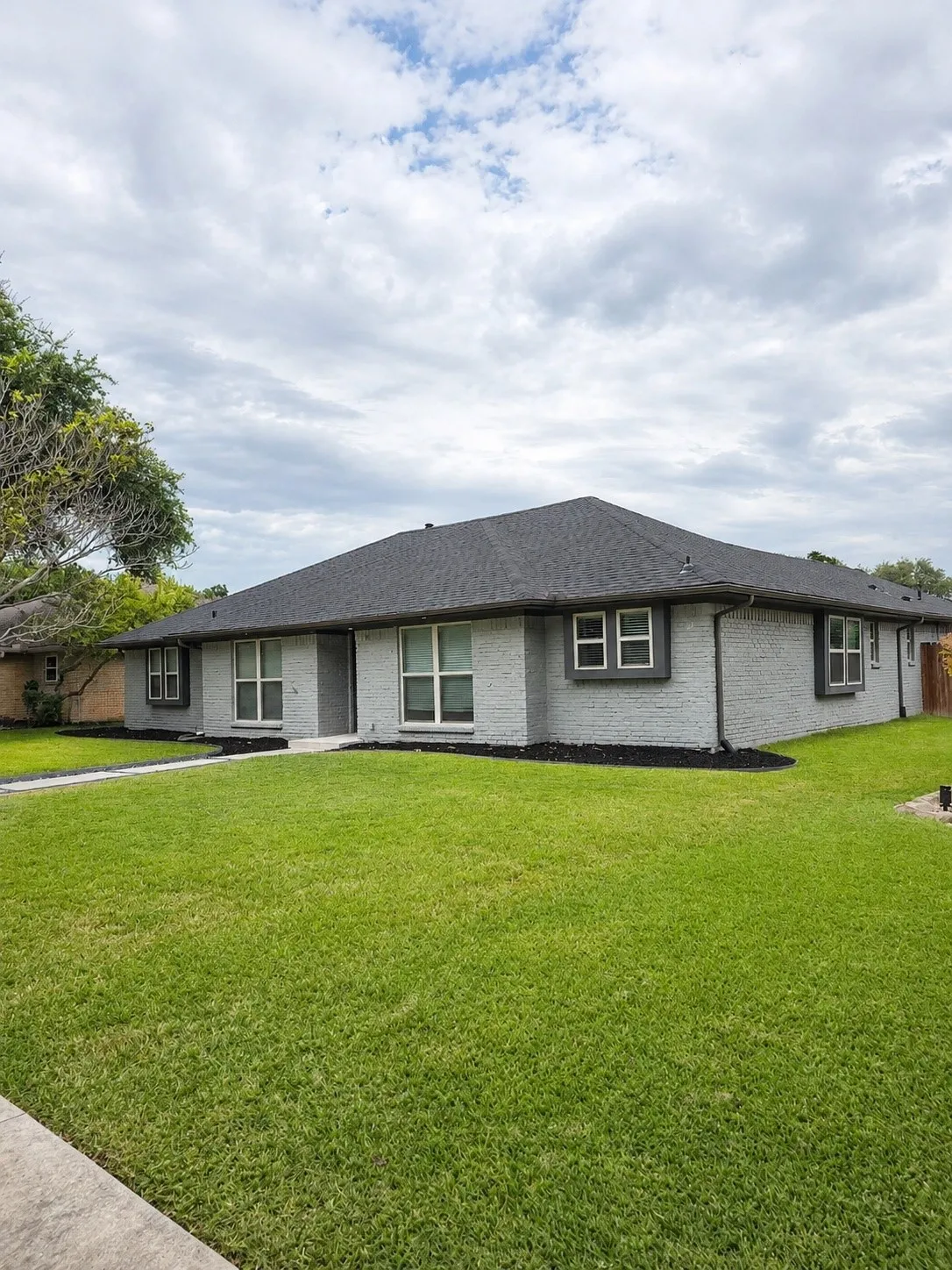 Modern single-story residence featuring a light brick exterior with dark architectural roofing, expansive front-facing windows, and a professionally manicured lawn with mature landscaping