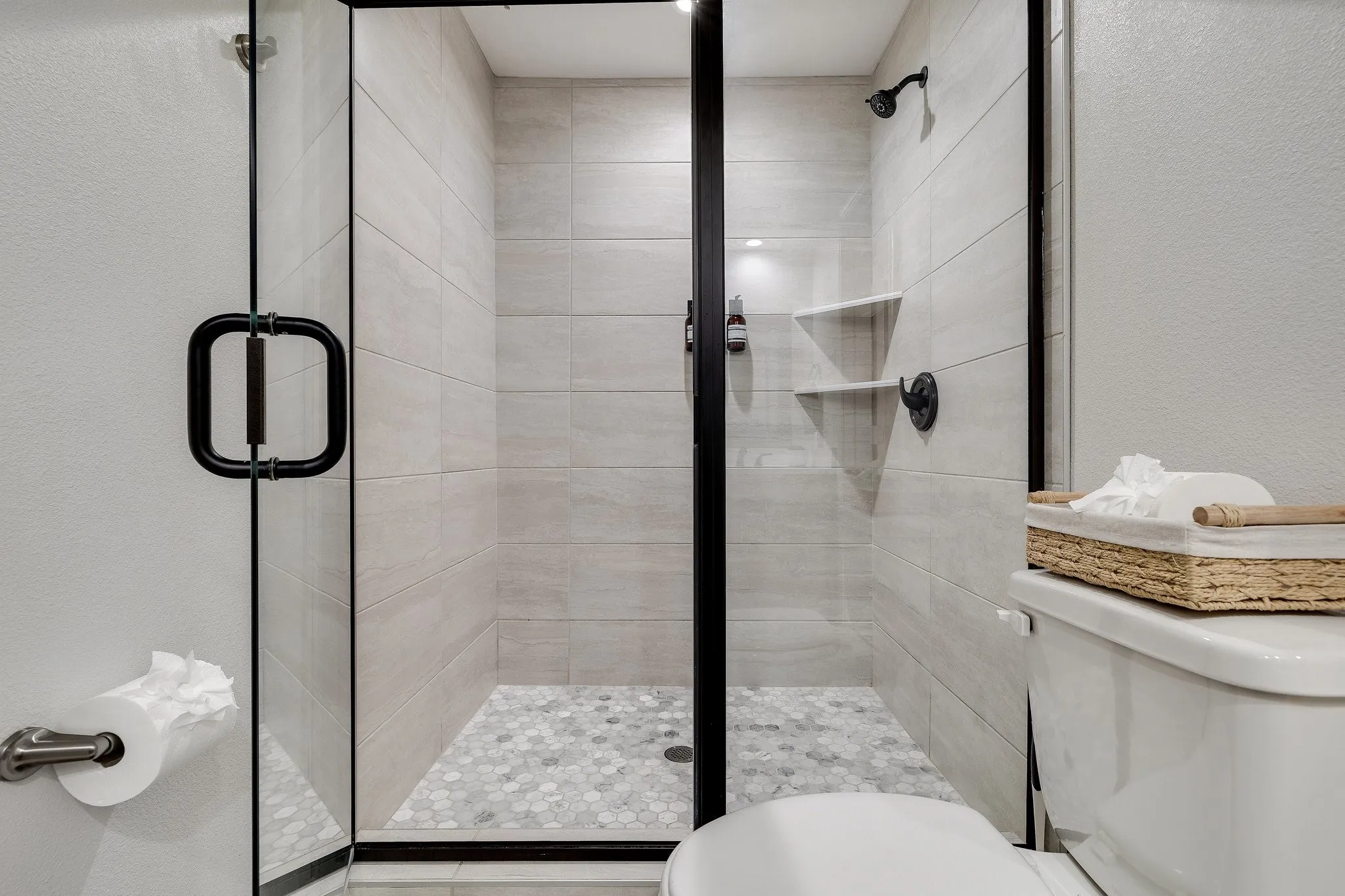 Glass-enclosed shower with matte black hardware, light-toned horizontal tile walls, mosaic tile shower pan, and built-in corner shelving