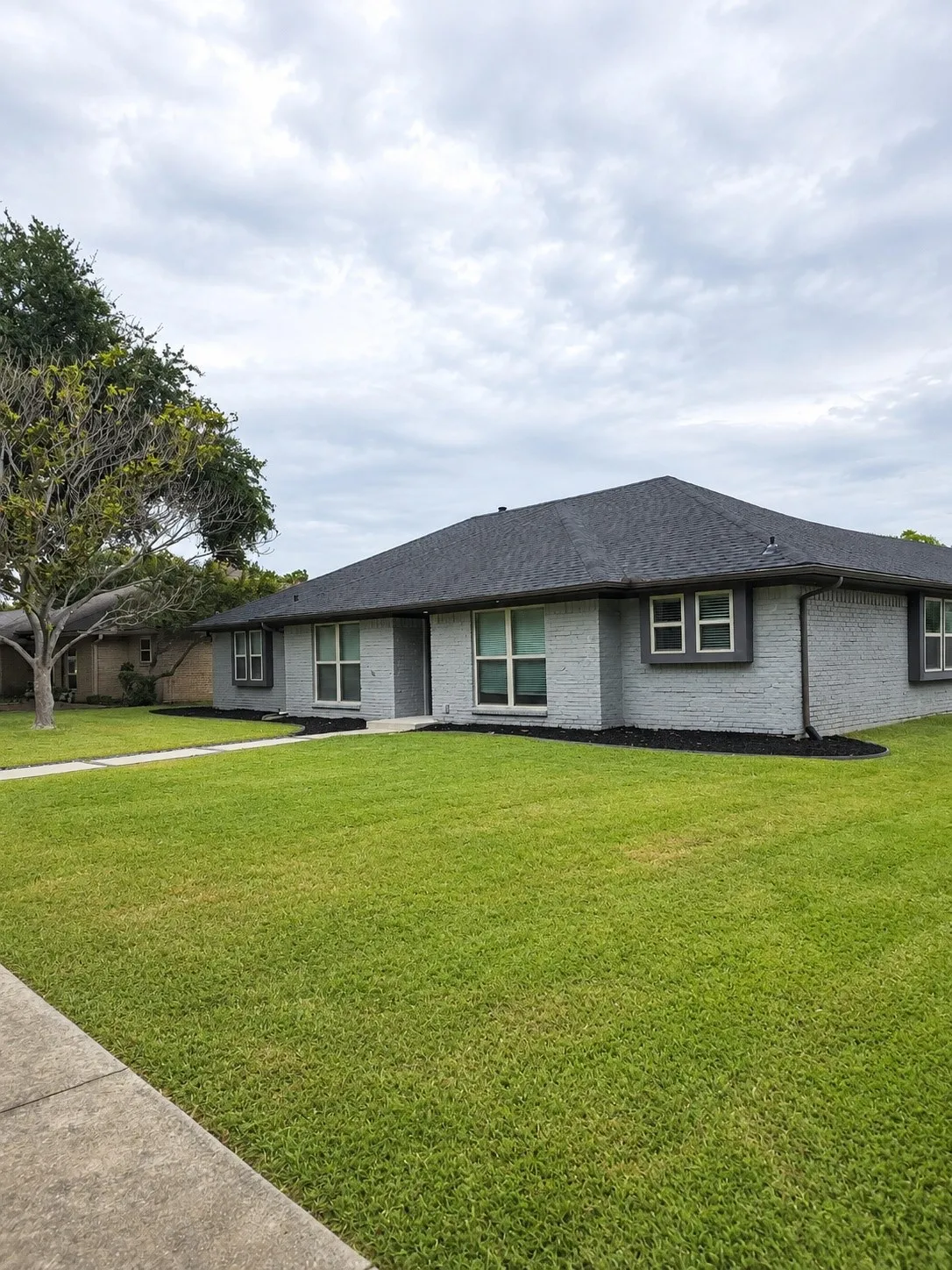 Gray brick exterior with dark trim, dark shingles, and a manicured lawn