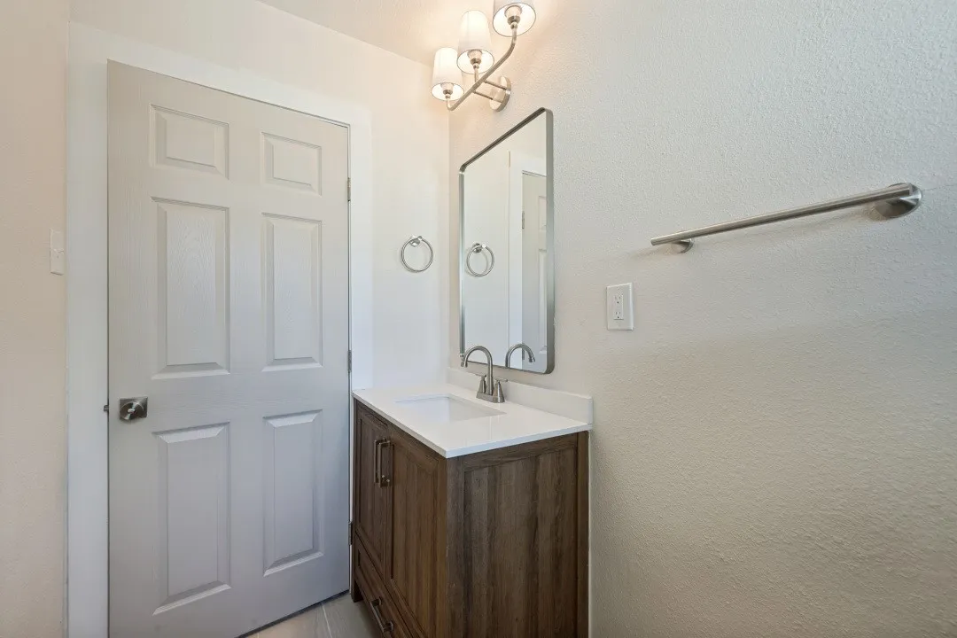 Refreshed vanity area with quartz-style countertop, wood-tone cabinetry, contemporary mirror, updated lighting, and coordinated brushed-metal fixtures.