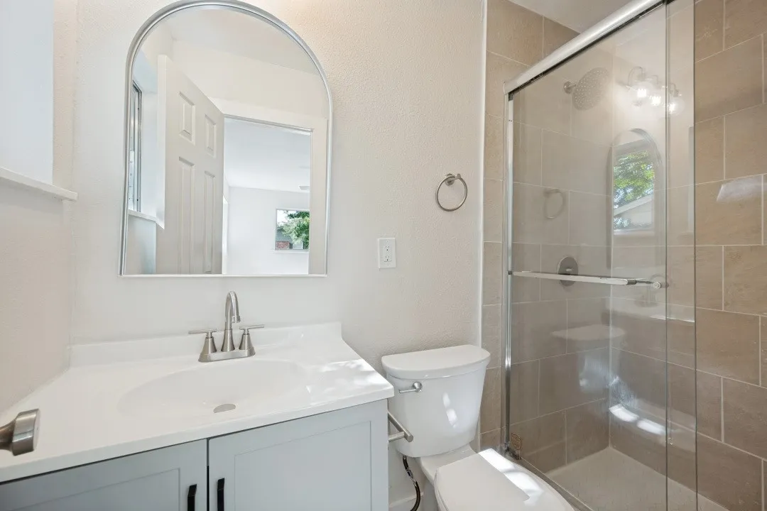 Renovated en-suite bath featuring a modern vanity, arched mirror, glass-enclosed tiled shower, rainfall-style shower head, and updated fixtures.