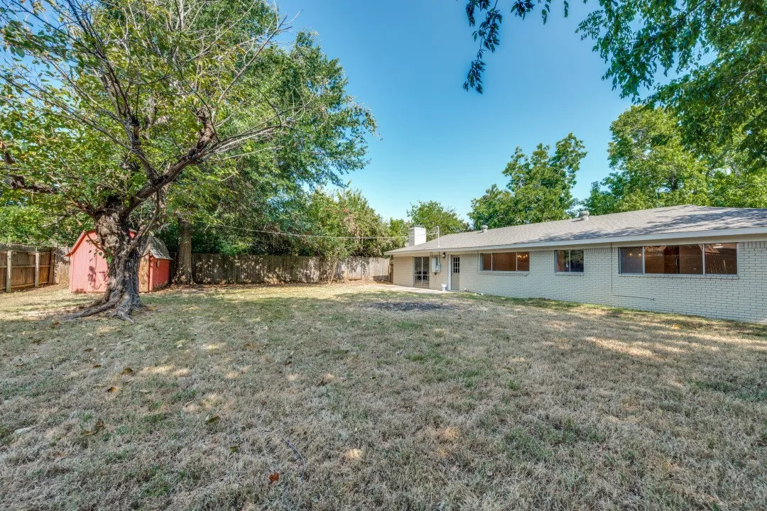 Spacious fenced backyard shaded by mature trees, with an expansive rear elevation, multiple windows, outdoor access, and a detached storage shed.