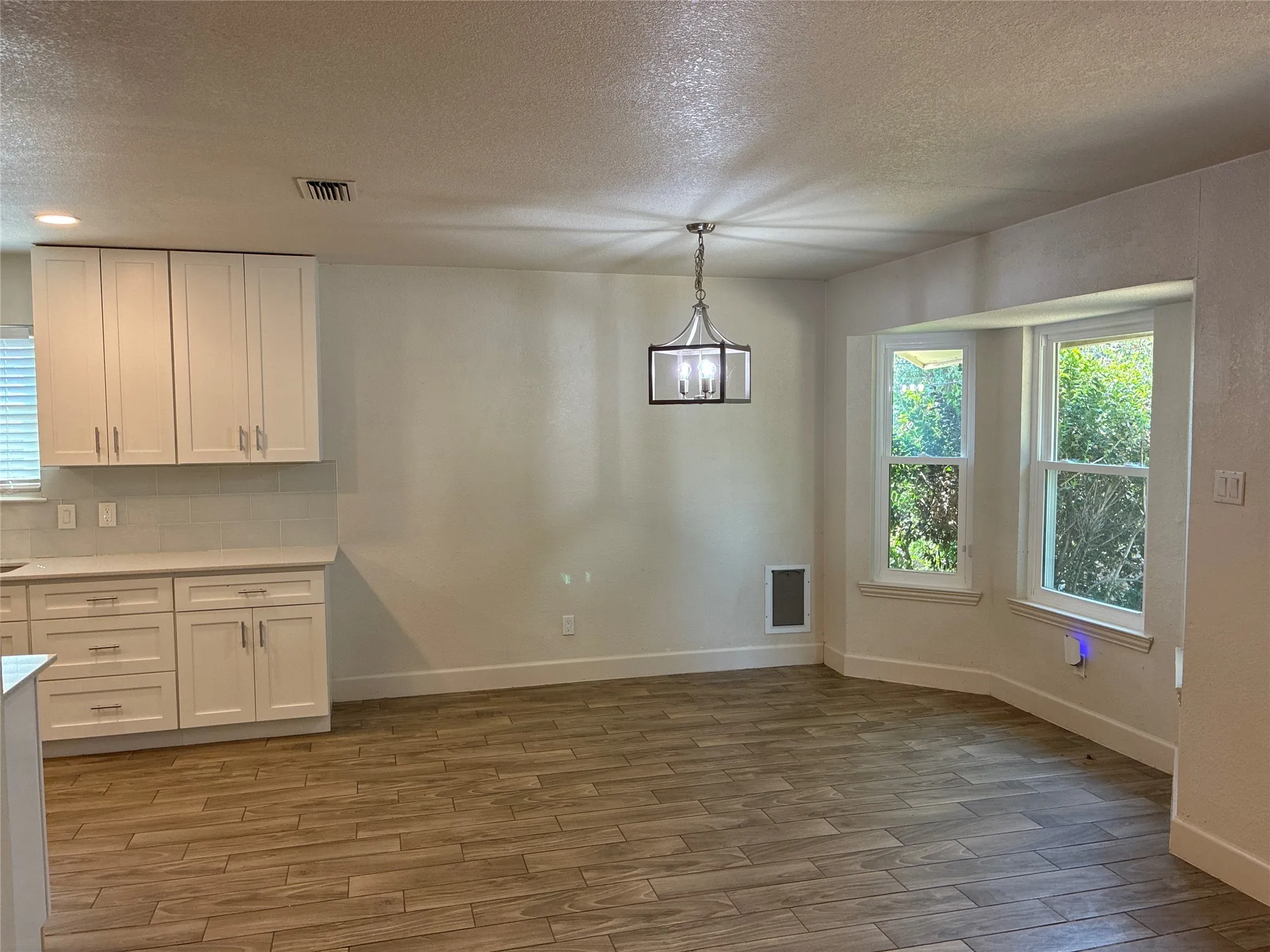 Bay window with white trim, wood-finish flooring, white cabinetry, tile backsplash, and a contemporary pendant light fixture